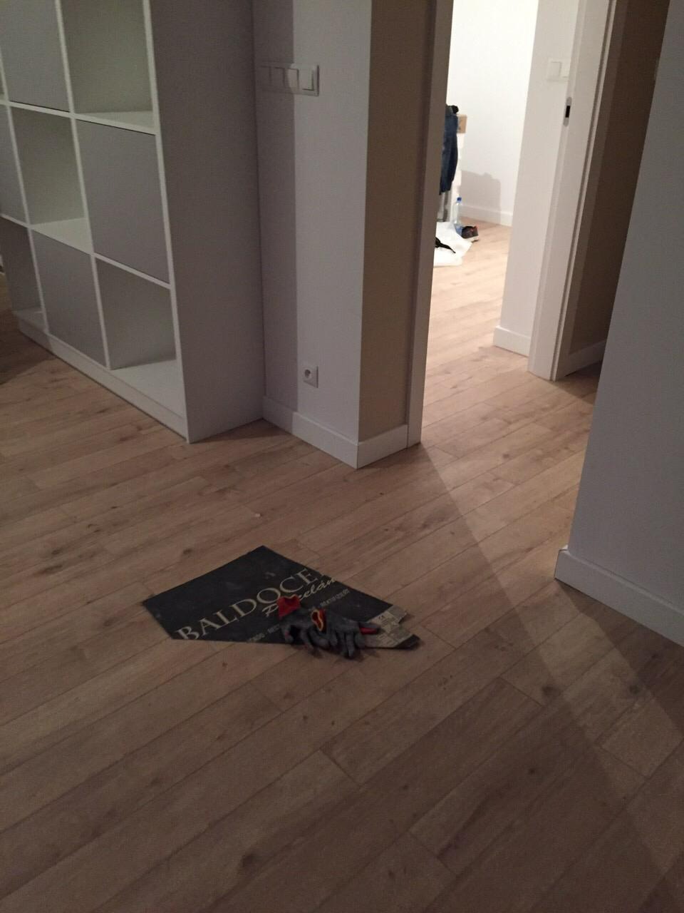 Jasne panele podłogowe w korytarzu z widoczną okładziną ochronną i rękawicami roboczymi, częściowo widoczny regał i otwarte drzwi do innego pomieszczenia.