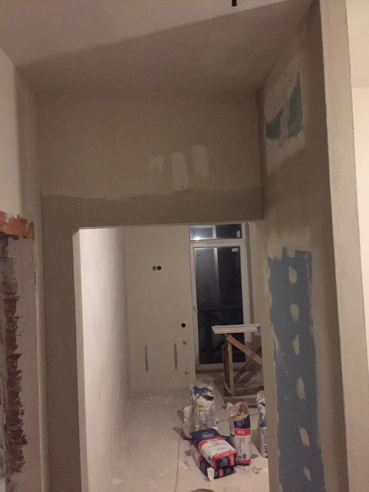 Wnętrze w trakcie prac wykończeniowych: częściowo wyszpachlowane ściany, worki z zaprawą na podłodze, widoczne ślady po wcześniejszych warstwach na ścianie przy futrynie.