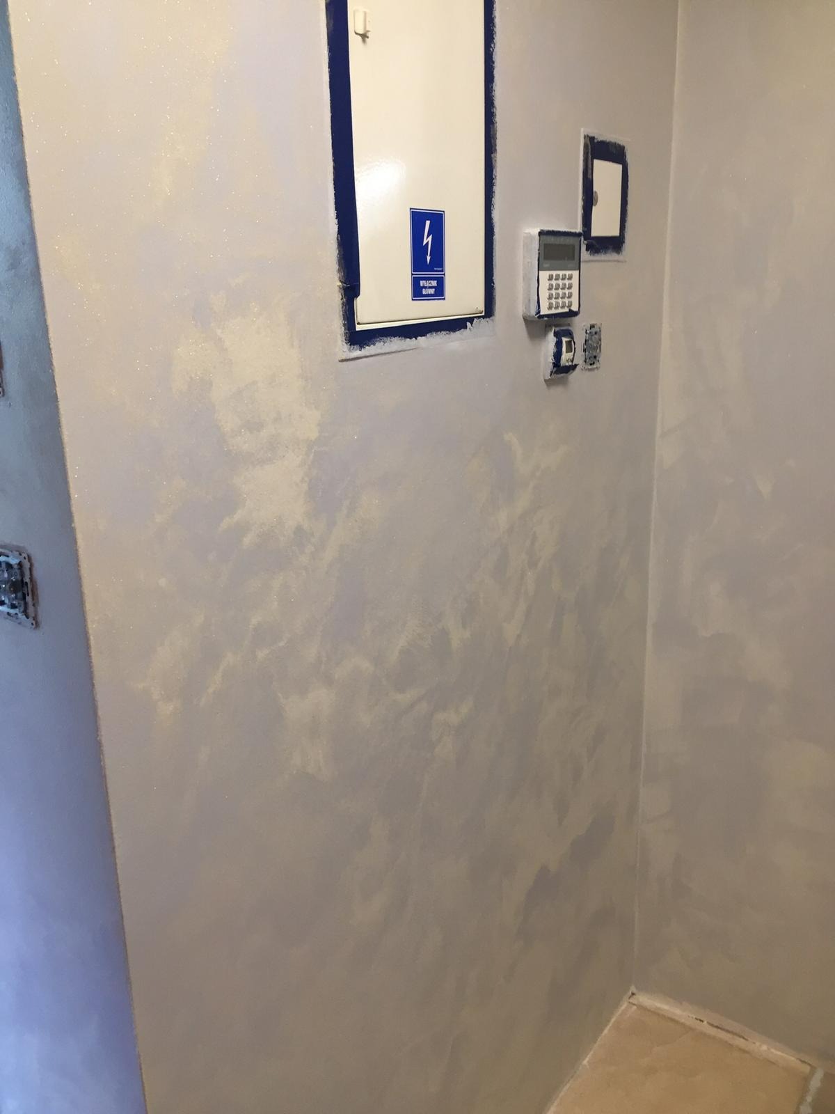Ściana w korytarzu z efektem dekoracyjnym Jeger, wykończona gładzią imitującą chmury, z widoczną skrzynką elektryczną i panelem alarmowym.