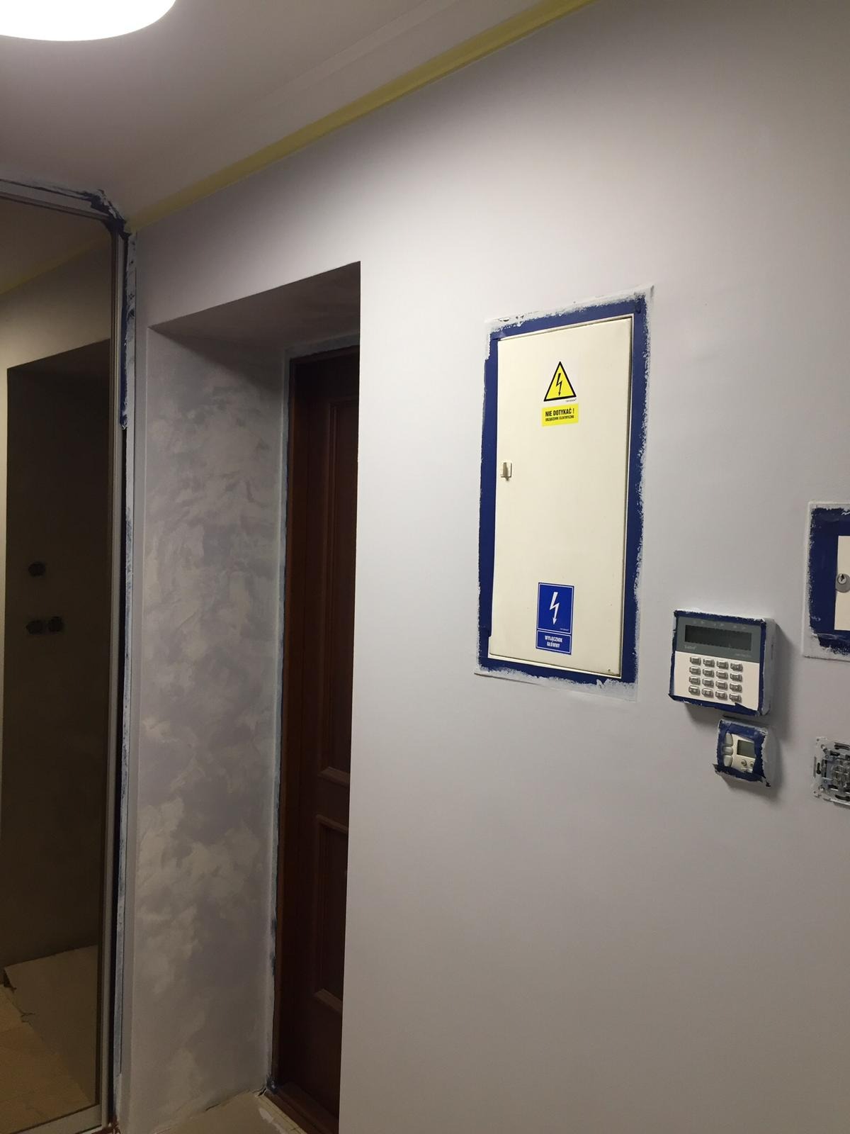 Świeżo pomalowana ściana w odcieniach szarości z zabezpieczonymi taśmą malarską elementami elektrycznymi, widoczna faktura dekoracyjna obok futryny drzwi.