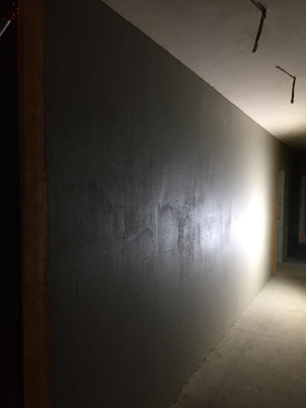 Klatka schodowa w trakcie prac: ściana pokryta szarą warstwą wykończeniową, widoczne niedoskonałości i nierówności podłoża, surowe oświetlenie korytarza.