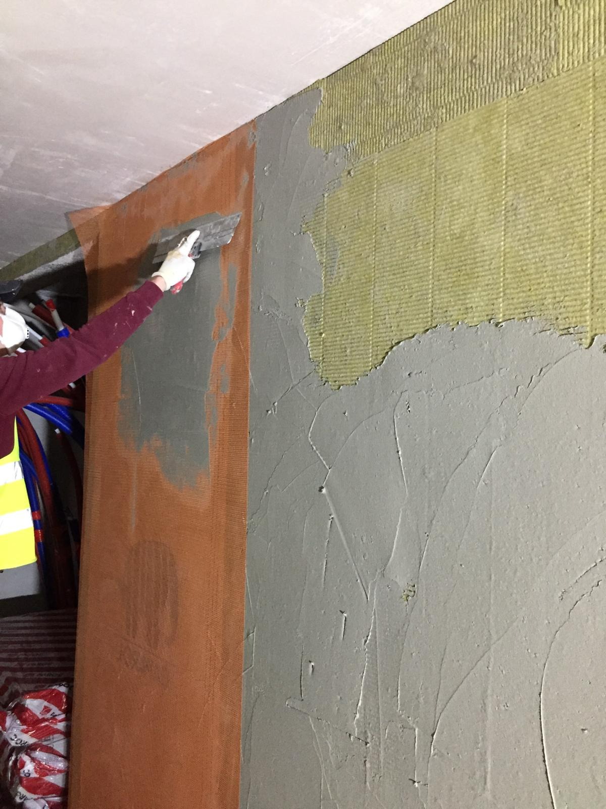 Nakładanie szarej masy szpachlowej na ścianę z widoczną izolacją i pomarańczową siatką wzmacniającą, pracownik w masce i rękawicach używa szpachelki.