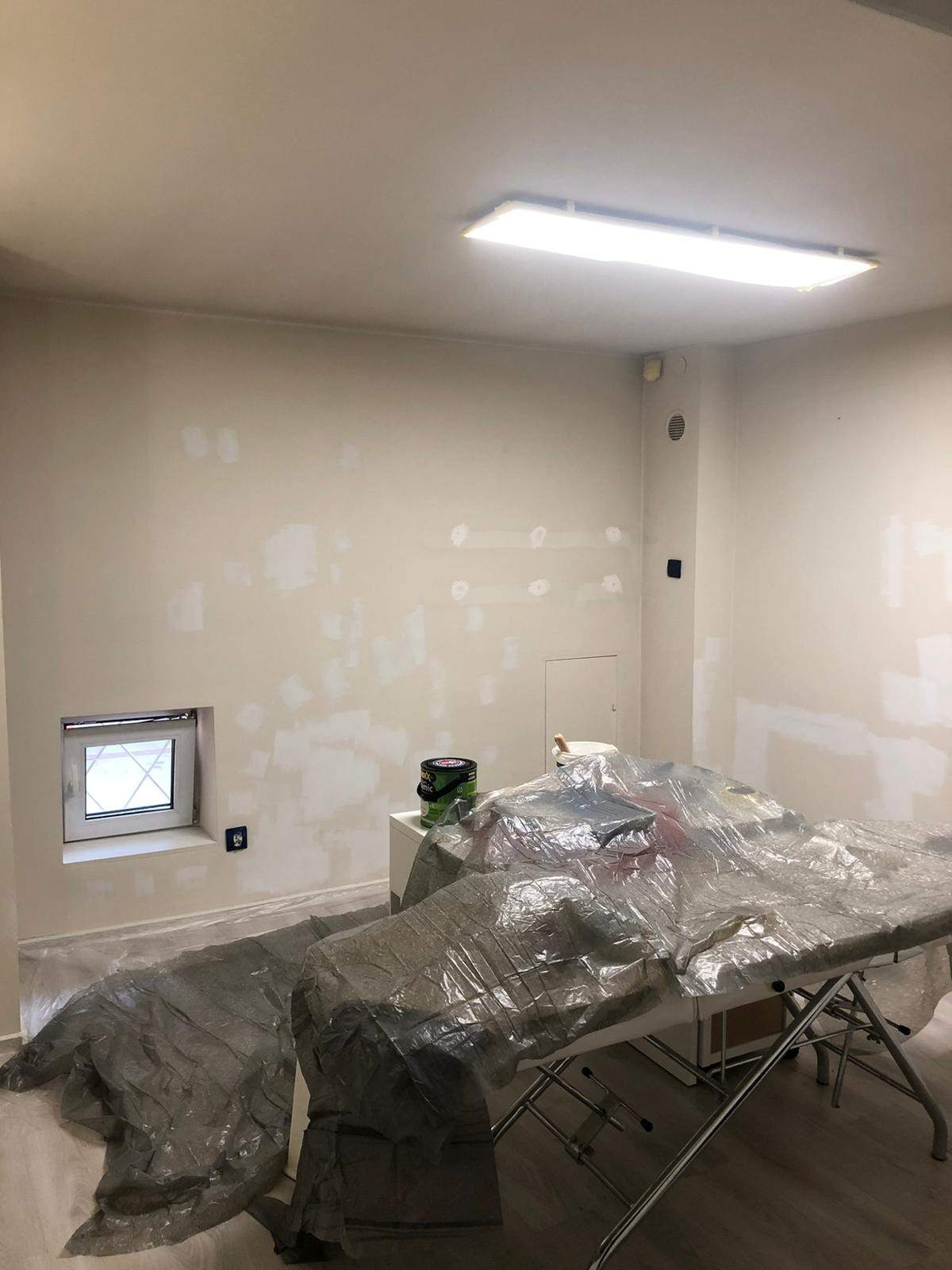 Przygotowanie gabinetu kosmetycznego do malowania: ściany po szpachlowaniu, zabezpieczone folią meble i podłoga, widoczne ślady po łataniu ubytków.