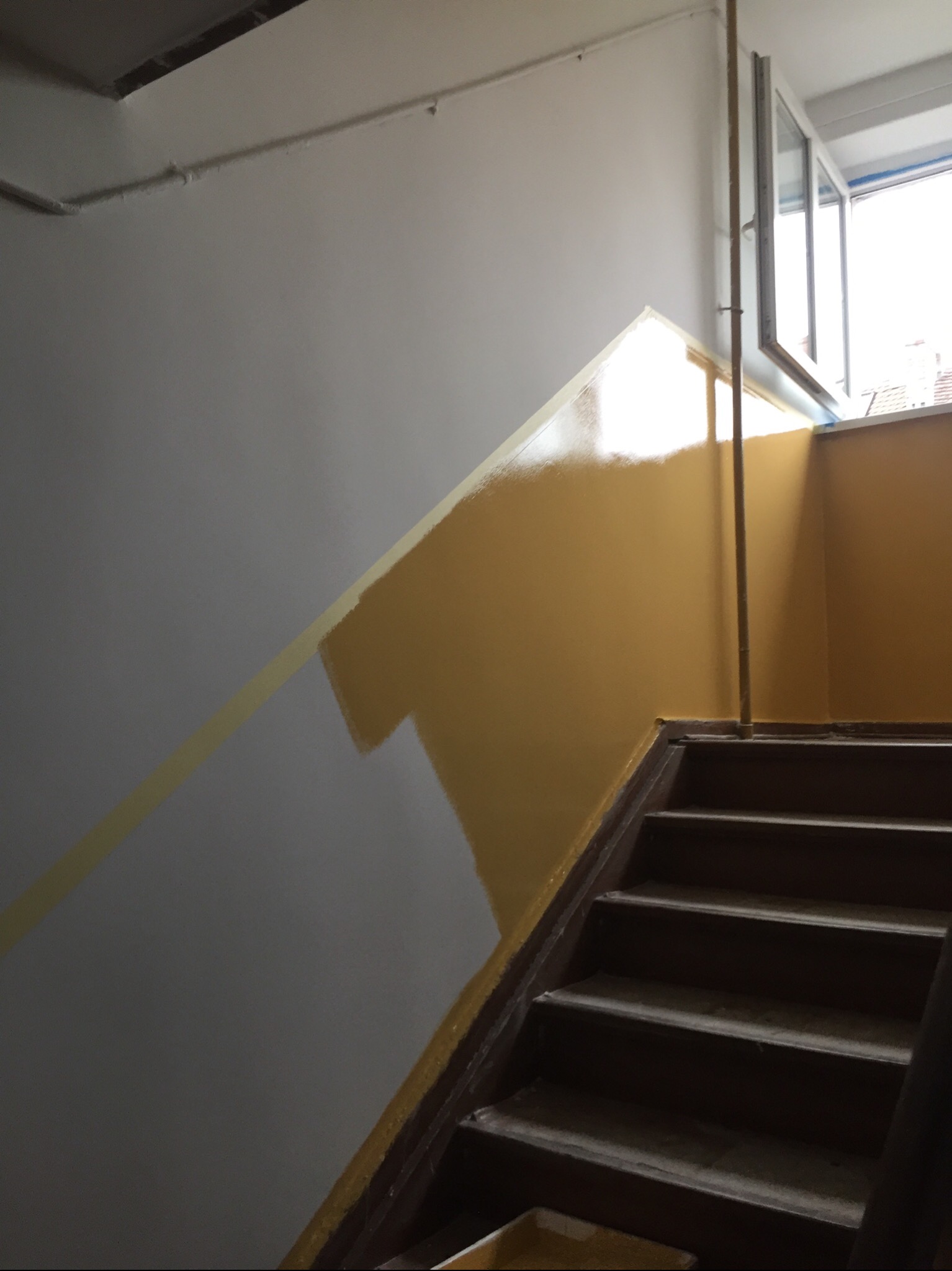 Malowanie klatki schodowej na żółto, z widoczną taśmą malarską oddzielającą pomalowaną część od szarej, z kilkoma stopniami schodów w dolnej części kadru.