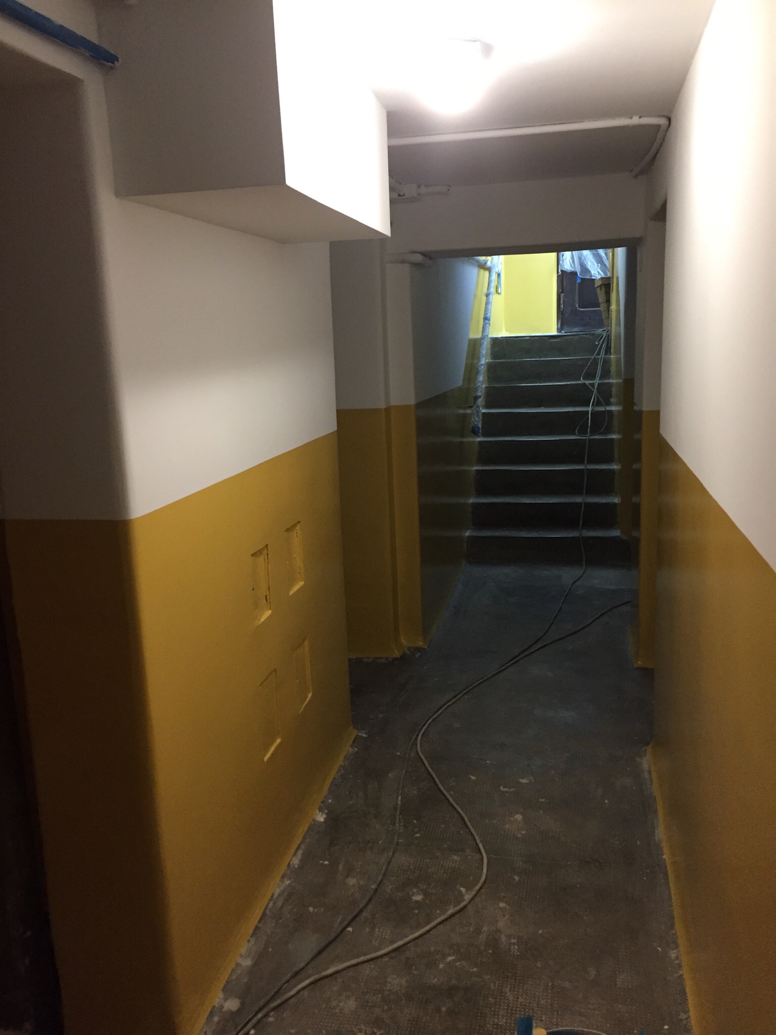 Korytarz w bloku po częściowym pomalowaniu ścian na biało i żółto, widoczne schody w głębi i kabel elektryczny na podłodze.