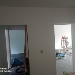Firma Ergo - Wnętrze w trakcie odświeżania w Gorzowie Wielkopolskim, widoczne otwarte drzwi, drabina malarska, puszki z farbą i zabezpieczona folią podłoga w kolejnym pomieszczeniu.