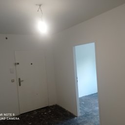 Firma Ergo - Wnętrze po świeżym malowaniu na biało, widoczne ślady farby na podłodze, surowa żarówka zwisająca z sufitu, drzwi wejściowe i otwarte drzwi do innego pomieszczenia.