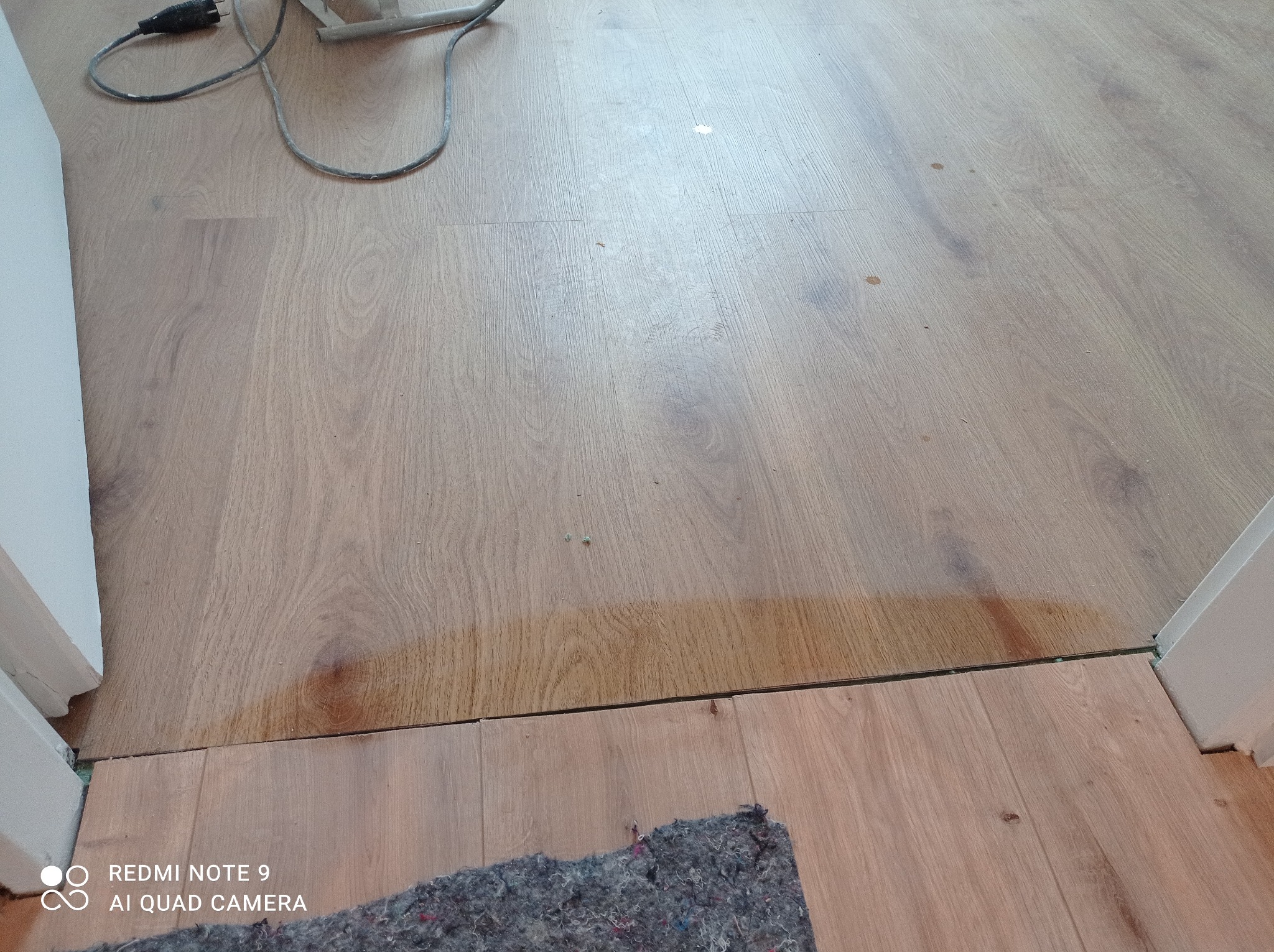 Fragment podłogi z paneli, widoczne niedociągnięcia w łączeniu przy progu, zarysowania i plamy na panelach, fragment dywanu i przewód od urządzenia.