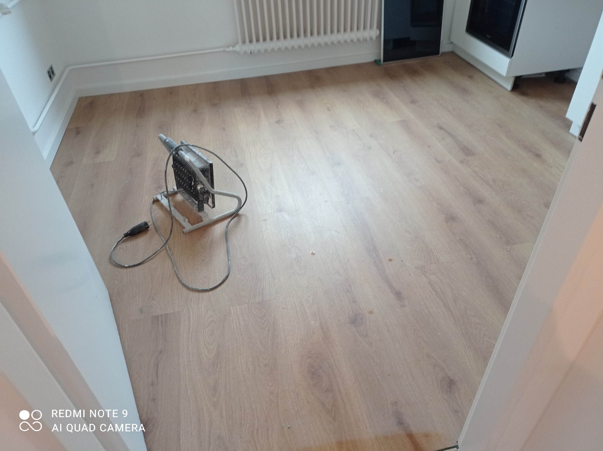 Świeżo ułożone panele podłogowe w jasnym odcieniu drewna, z widocznym grzejnikiem i fragmentem białej zabudowy kuchennej, oświetlone lampą halogenową na statywie.