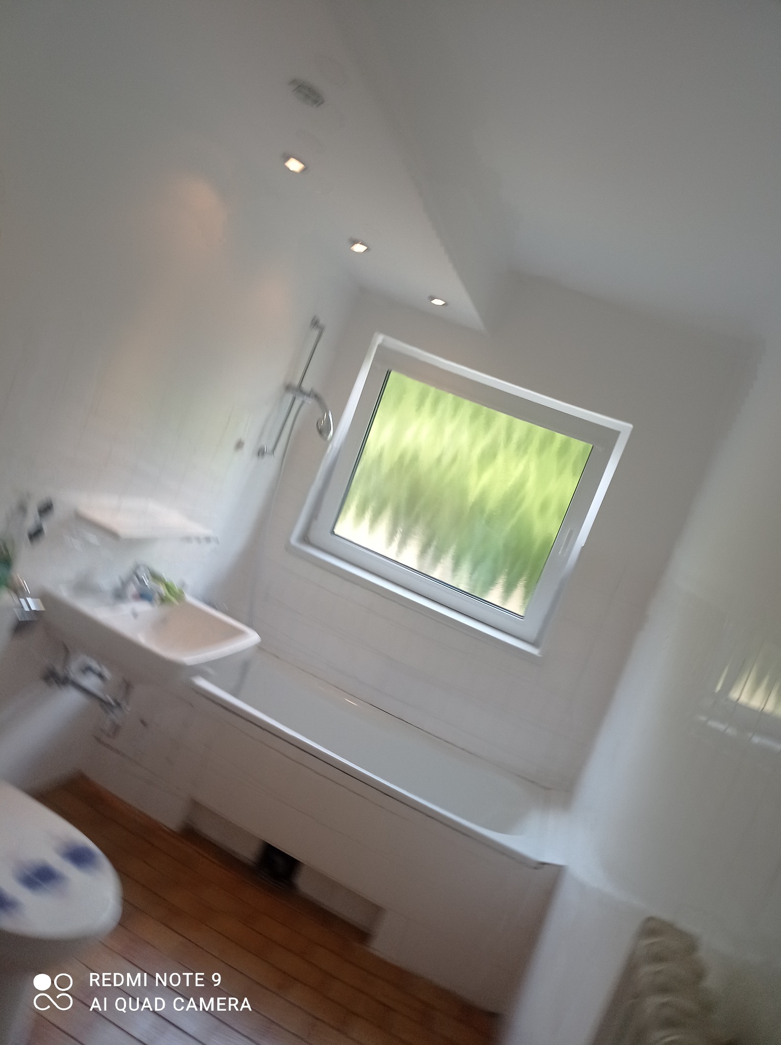 Jasna łazienka z drewnianą podłogą, białą armaturą, oknem z widokiem na zieleń i oświetleniem punktowym w suficie.