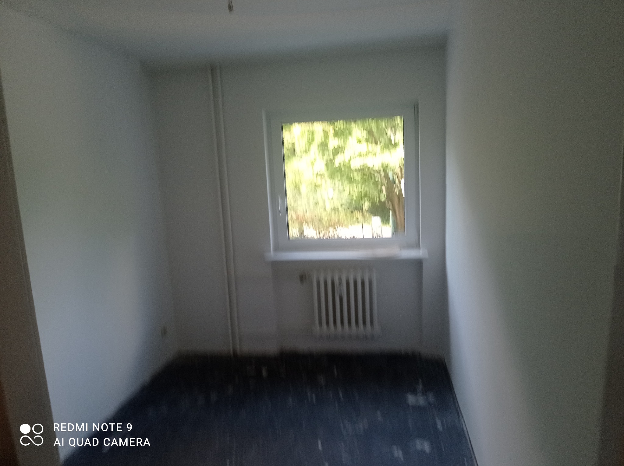 Pusty pokój z pomalowanymi na biało ścianami, grzejnikiem pod oknem i ciemną podłogą w trakcie przygotowania do dalszych prac wykończeniowych.