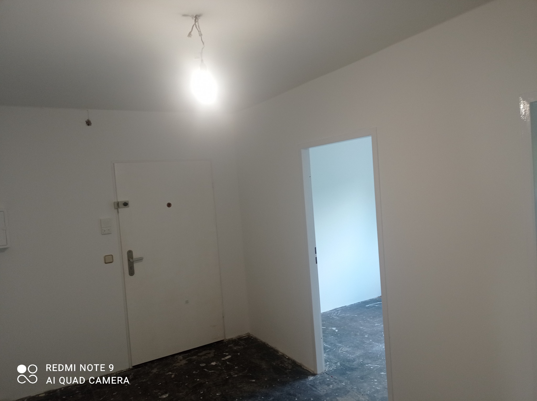 Wnętrze po świeżym malowaniu na biało, widoczne ślady farby na podłodze, surowa żarówka zwisająca z sufitu, drzwi wejściowe i otwarte drzwi do innego pomieszczenia.
