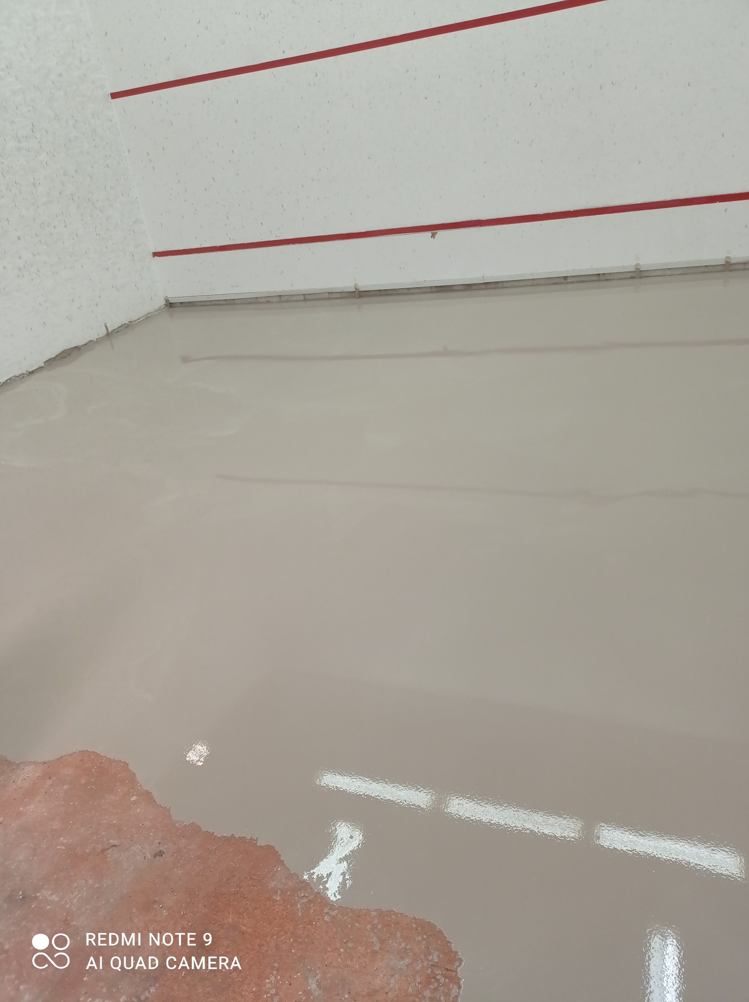 Świeżo wylana, samopoziomująca wylewka podłogowa w jasnym odcieniu szarości, z widocznym fragmentem starego, czerwonego podłoża przy krawędzi i czerwonymi liniami na ścianie w tle.