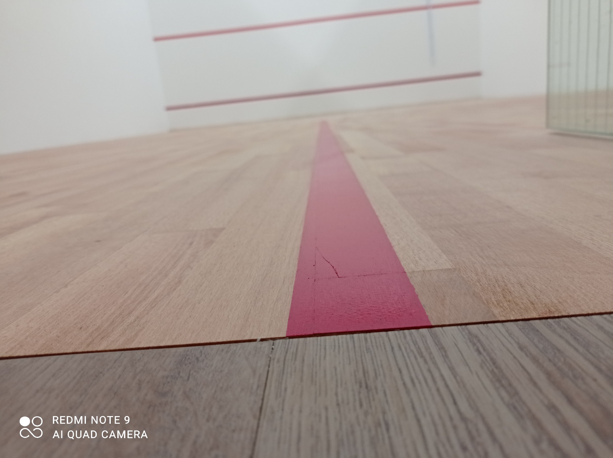 Parkietowa podłoga w hali sportowej ze świeżo wymalowaną czerwoną linią, widoczne niedoskonałości i pęknięcia farby.