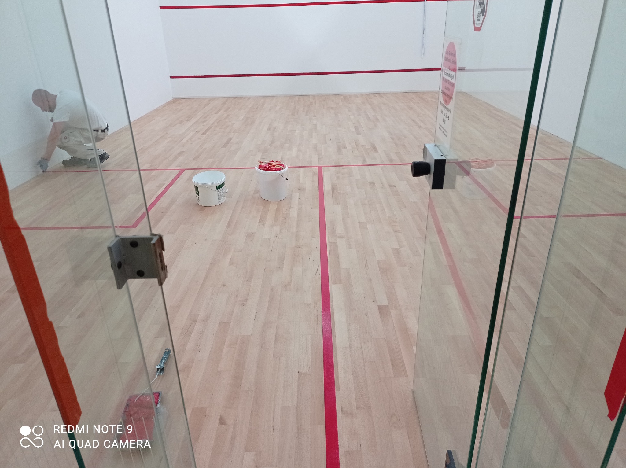 Pracownik maluje dolną część ściany wewnątrz kortu do squasha, widoczne dwa wiadra z farbą, podłoga w jasnym drewnie z czerwonymi liniami