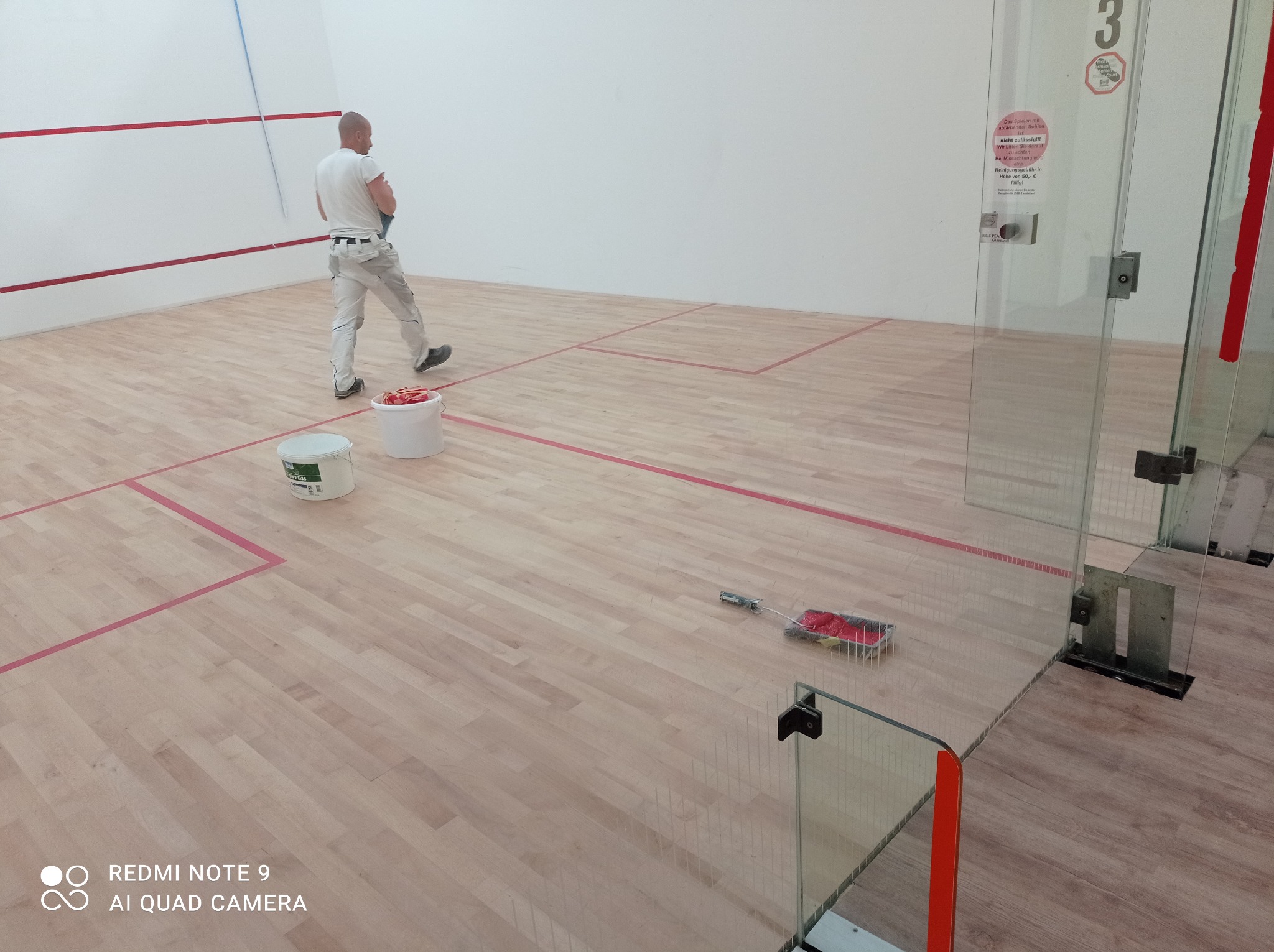 Mężczyzna w białej koszulce i spodniach maluje ściany w pomieszczeniu sportowym, widoczne dwa wiadra z farbą, wałek i pędzel, podłoga parkietowa z czerwonymi liniami.