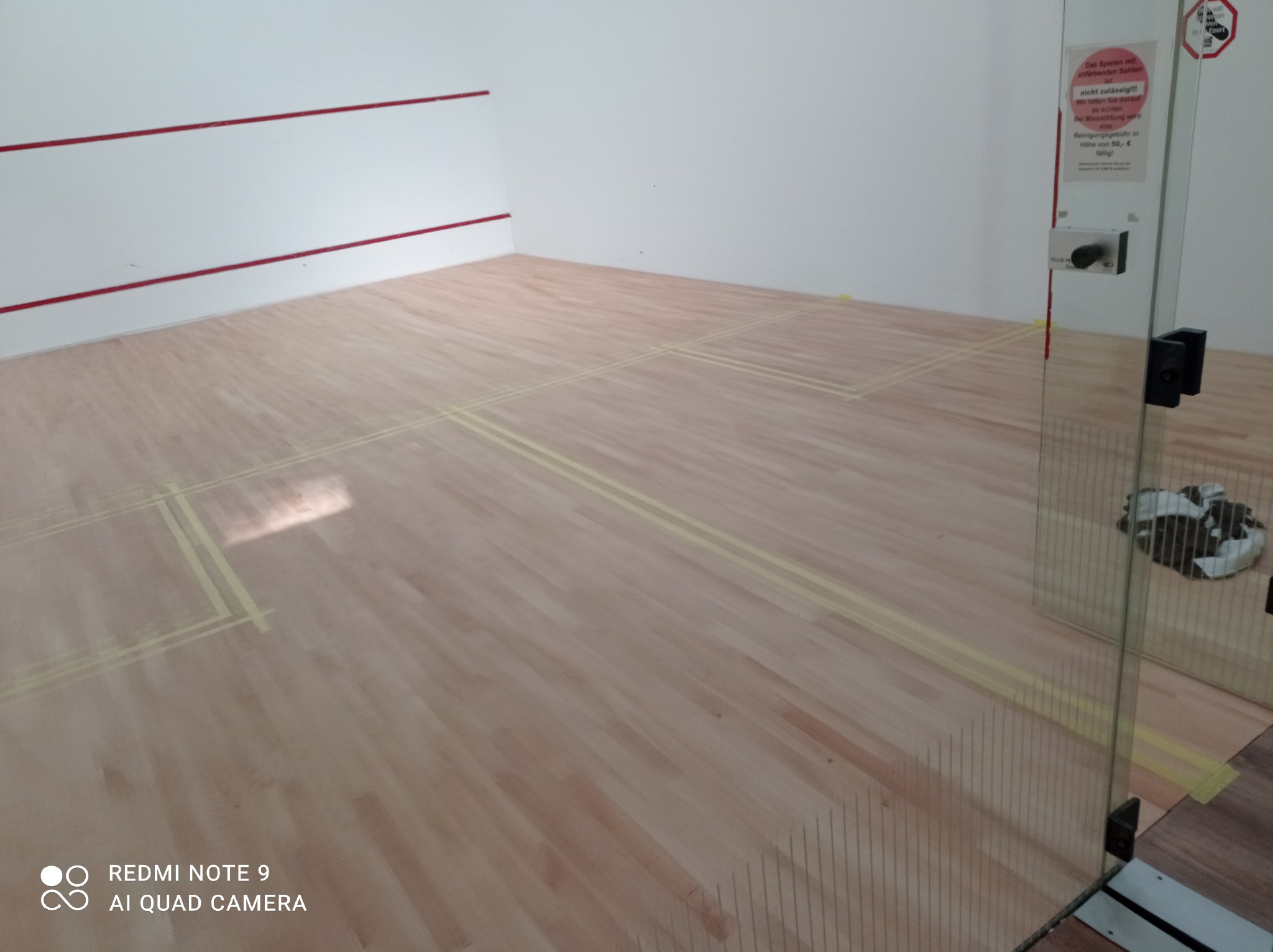 Sala do squasha z nowym parkietem z jasnego drewna i żółtymi liniami, widoczne drzwi szklane z informacją w języku niemieckim.
