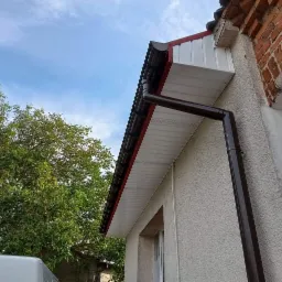 Zamontowana, brązowa rynna spustowa na elewacji budynku z fragmentem dachu krytego blachodachówką, widok z dołu na tle błękitnego nieba i drzewa.