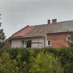Dom z częściowo wymienionym dachem, widoczne krokwie i nowe czerwone dach&oacute;wki obok starego pokrycia z eternitu, drabina i rusztowanie przy ścianie budynku.