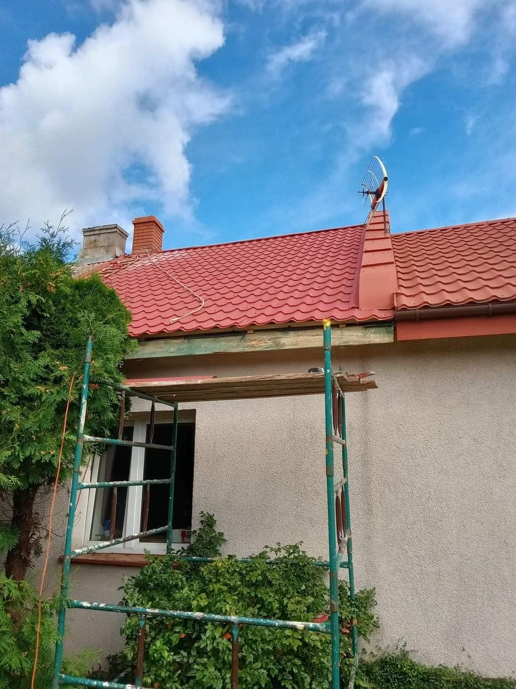 Czerwony dach z metalowej dachówki, antena satelitarna na kominie, rusztowanie przy ścianie budynku, błękitne niebo z chmurami w tle.