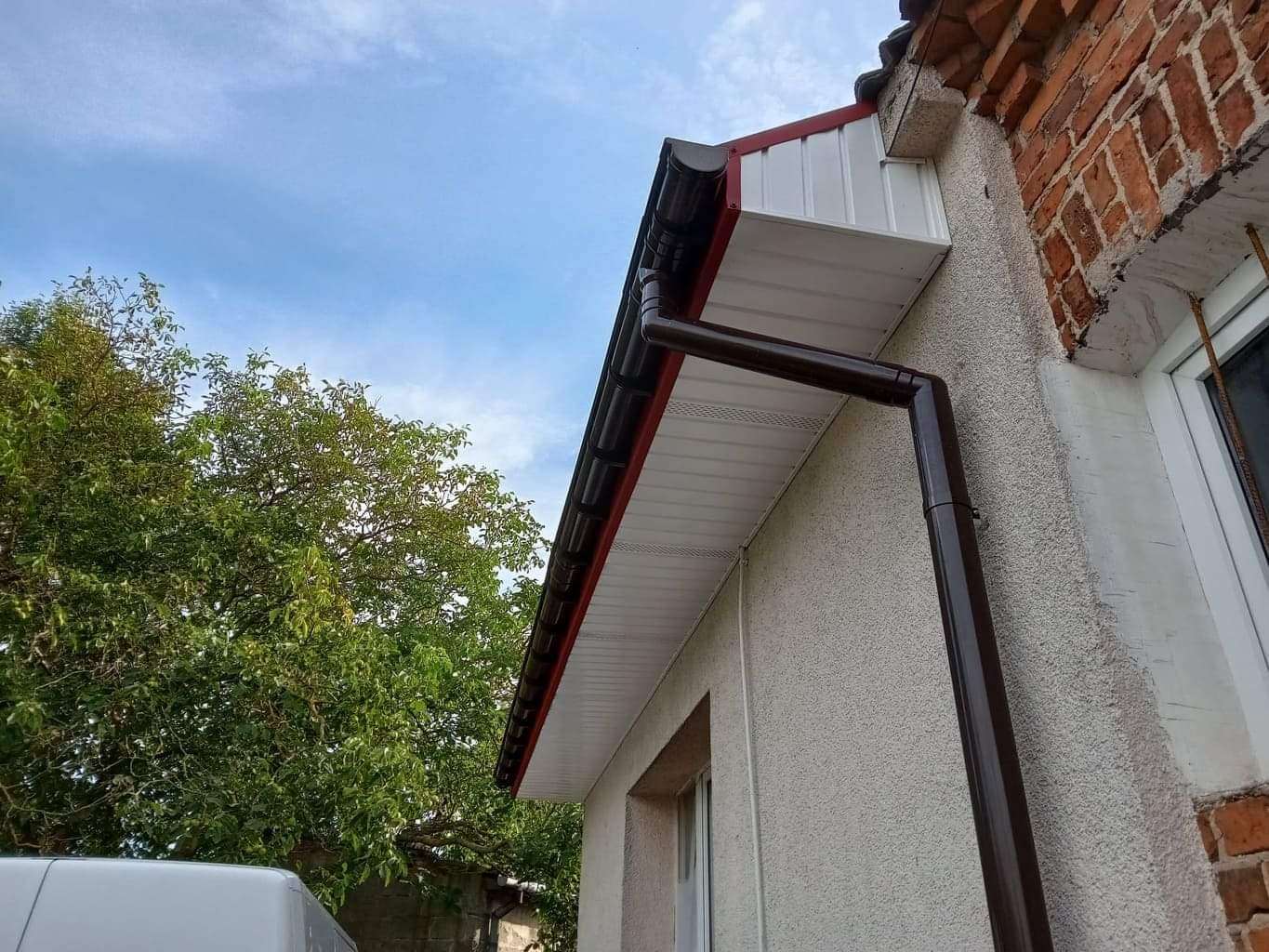 Zamontowana, brązowa rynna spustowa na elewacji budynku z fragmentem dachu krytego blachodachówką, widok z dołu na tle błękitnego nieba i drzewa.