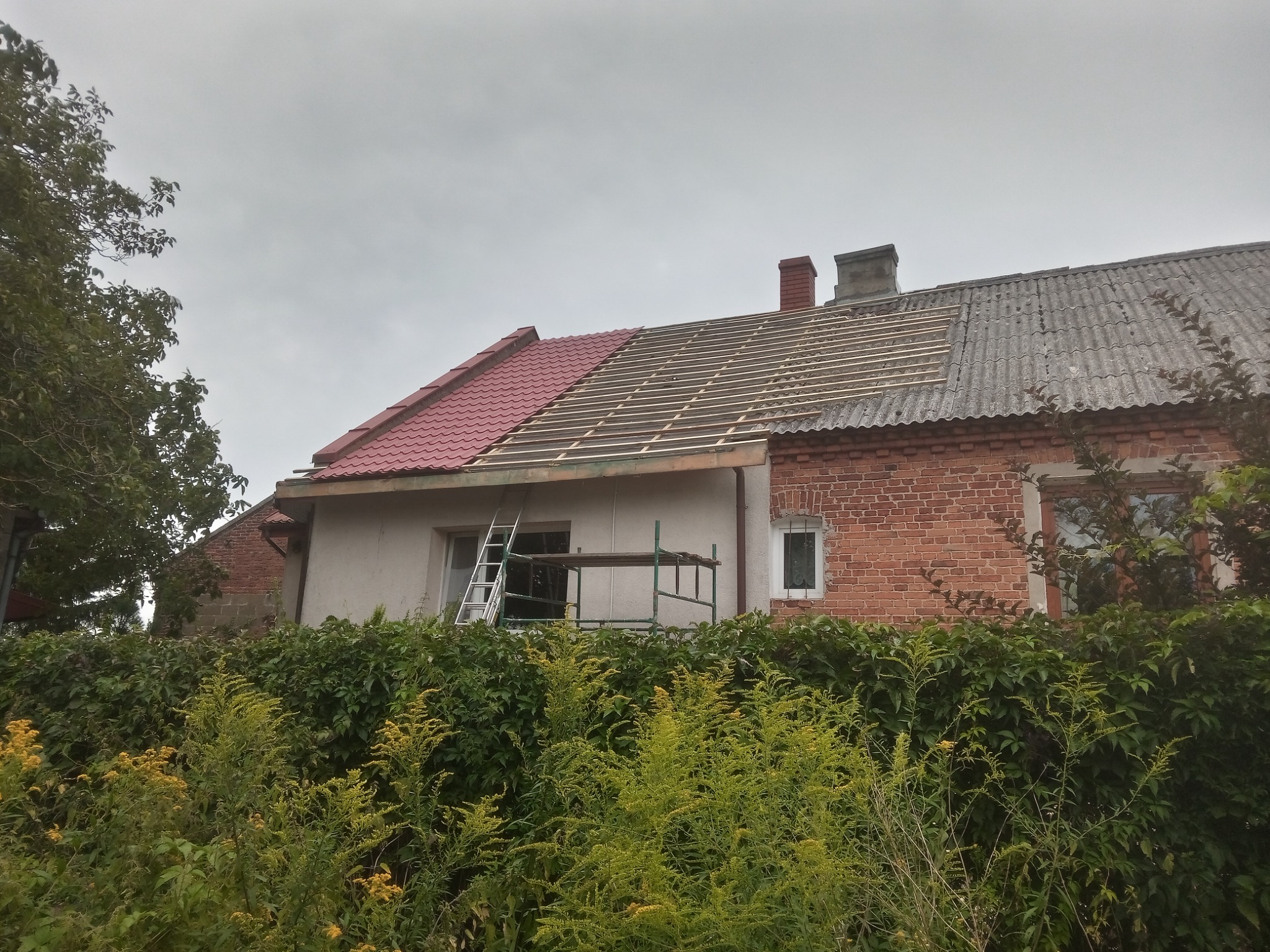 Dom z częściowo wymienionym dachem, widoczne krokwie i nowe czerwone dachówki obok starego pokrycia z eternitu, drabina i rusztowanie przy ścianie budynku.