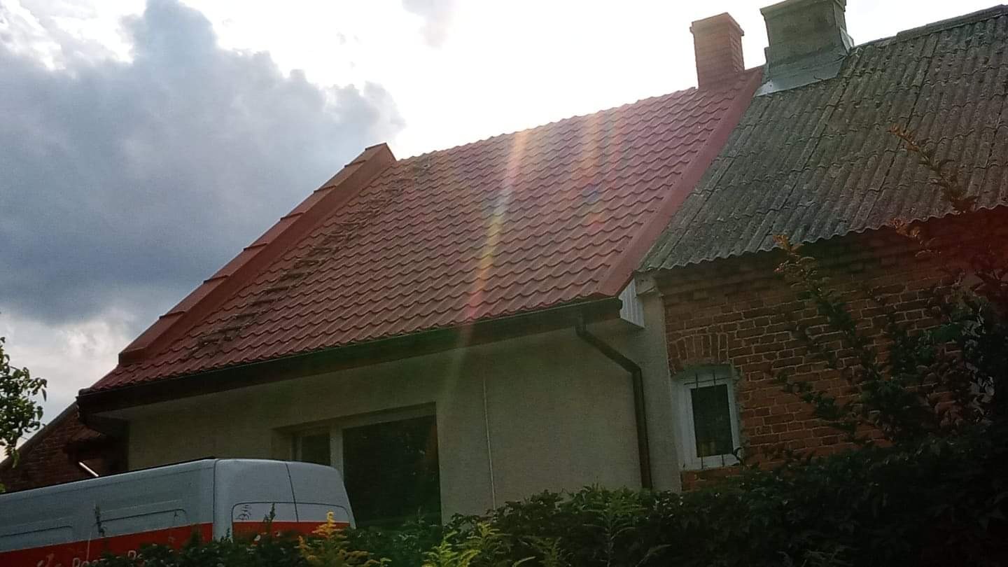 Dom z nową, czerwoną dachówką blachodachówkową, widoczne ślady zabrudzeń na powierzchni dachu, obok starszy budynek z szarą dachówką eternitową i ceglaną elewacją, na pierwszym planie krzewy...