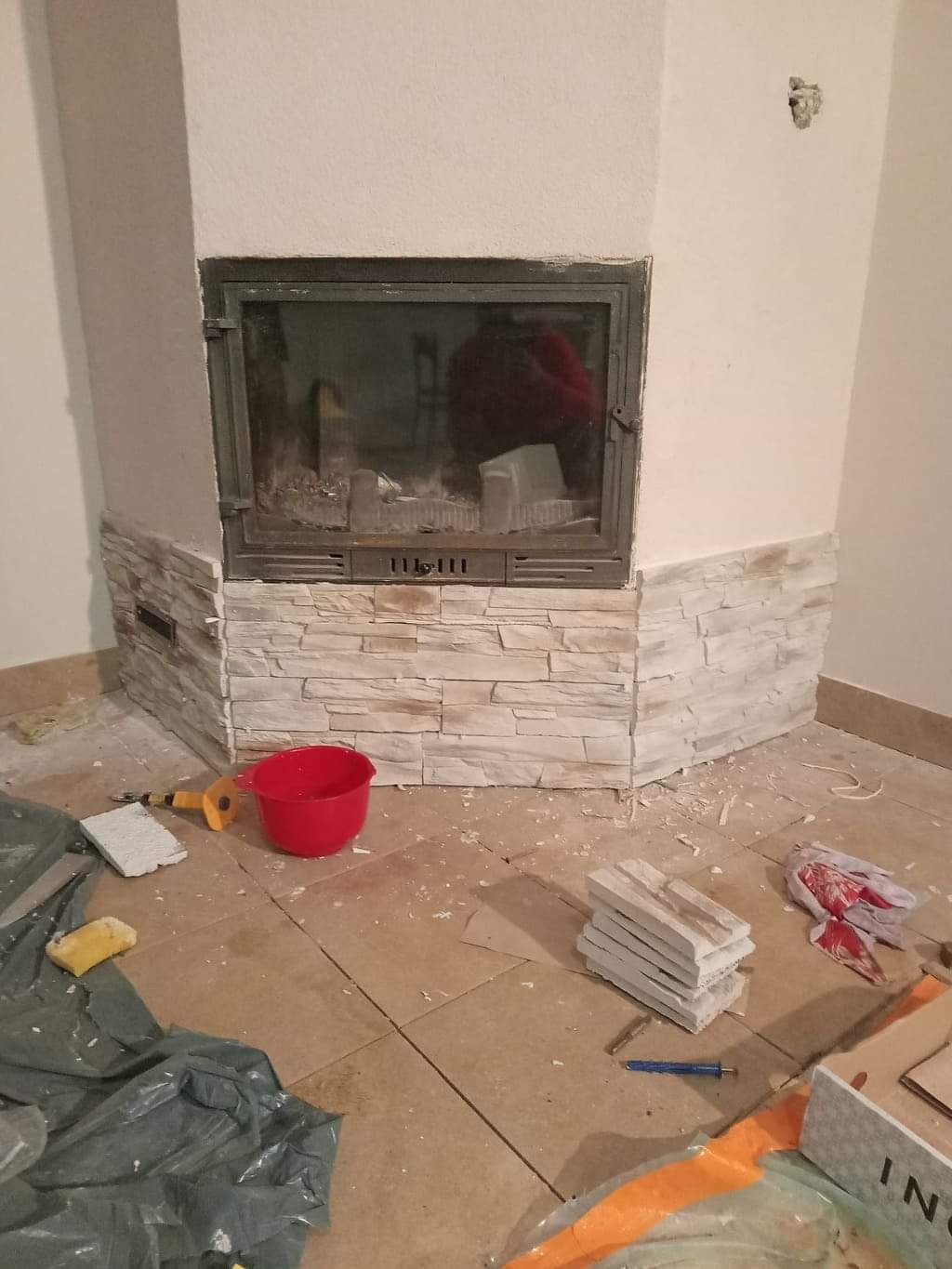 Wnętrze z kominkiem w trakcie obkładania dekoracyjnym kamieniem elewacyjnym, widoczne narzędzia i materiały budowlane, czerwone wiadro, fragmenty kamienia na podłodze.