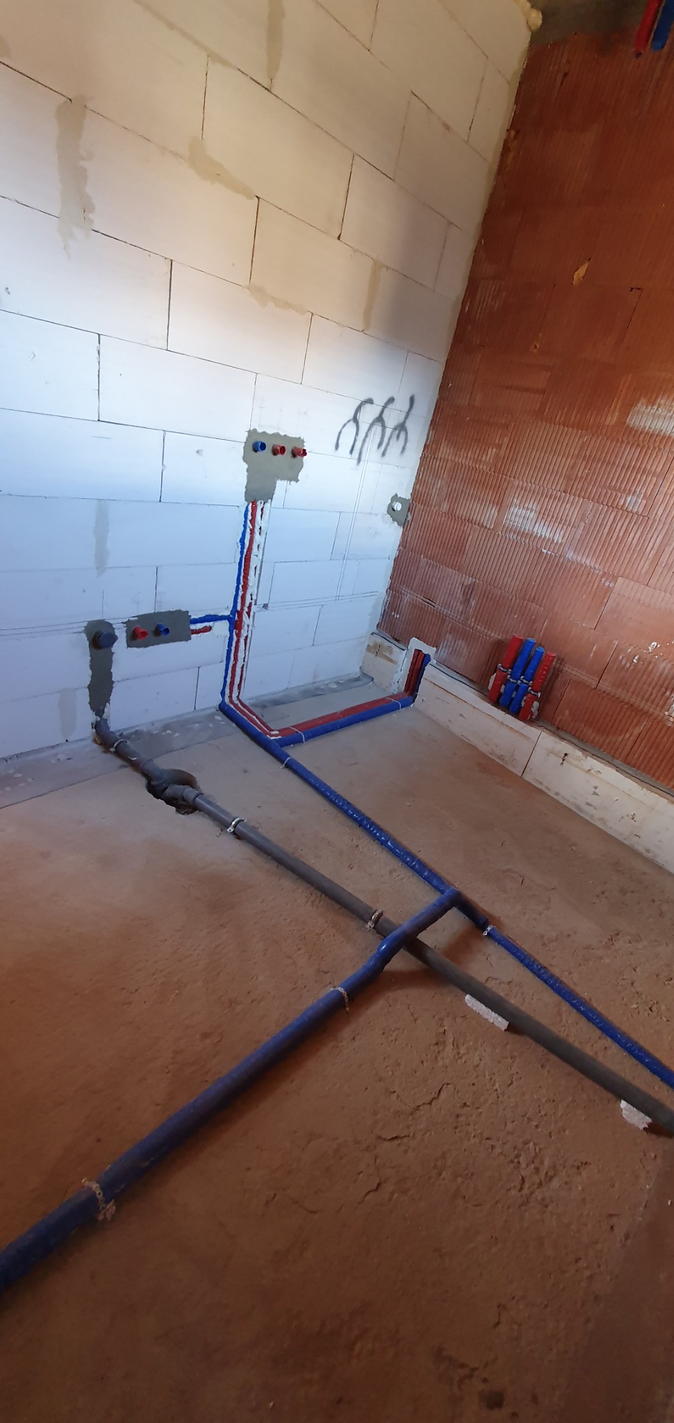 Wnętrze w trakcie prac instalacyjnych; widoczne rozprowadzone rury instalacji wodnej (czerwone i niebieskie) na ścianach z betonu komórkowego oraz szare rury kanalizacyjne w podłodze.