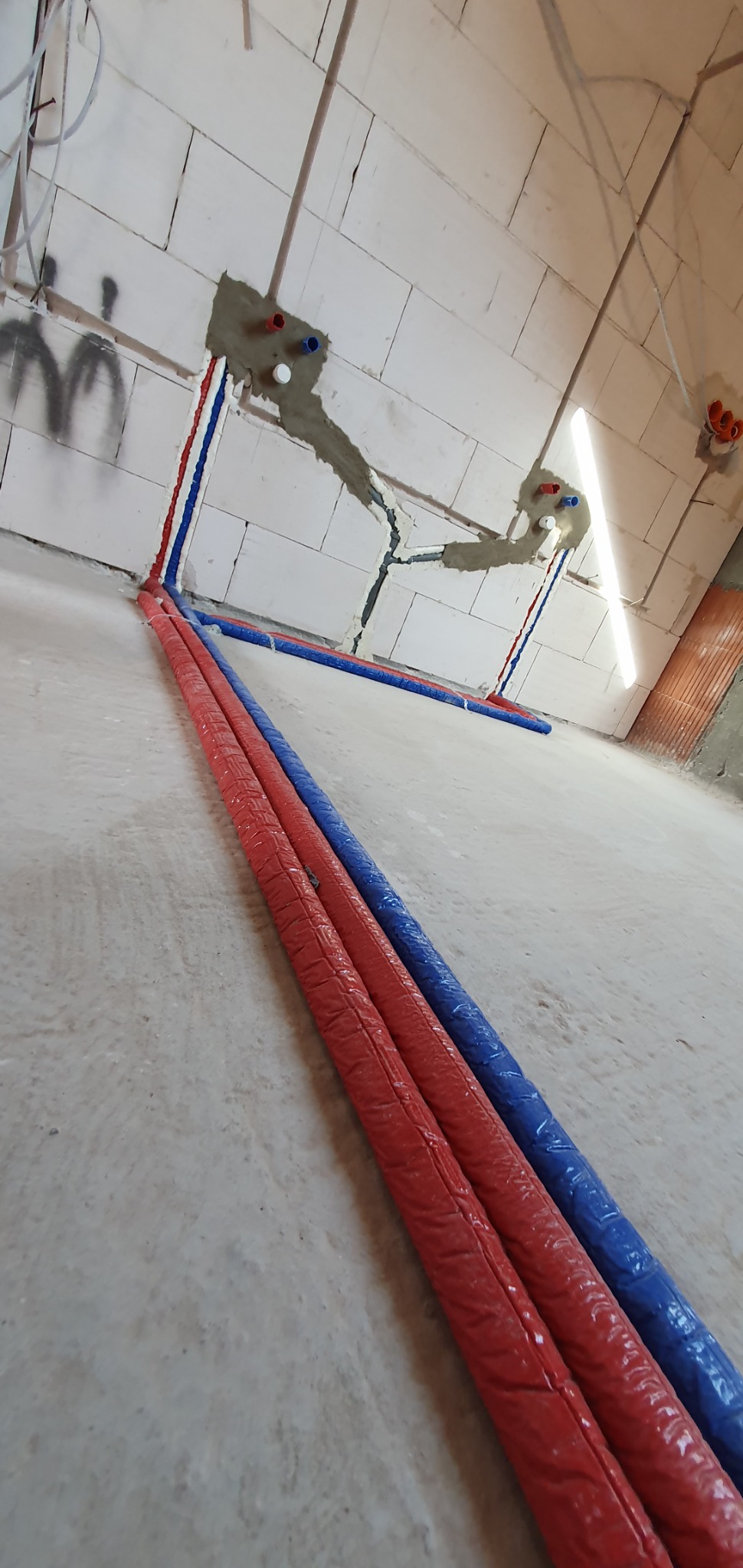 Nowe, izolowane rury hydrauliczne w kolorze czerwonym i niebieskim, prowadzone po podłodze i w ścianie z betonu komórkowego, z widocznymi punktami podłączeń.