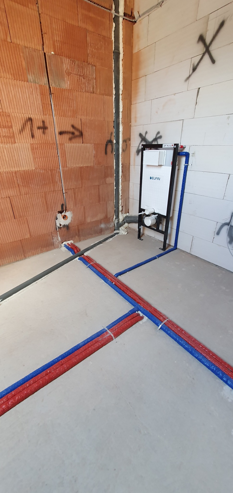 Surowe wnętrze z instalacją podtynkową WC marki Delfin, rurami grzewczymi w izolacji czerwono-niebieskiej krzyżującymi się na posadzce oraz pionami instalacyjnymi uszczelnionymi pianką.