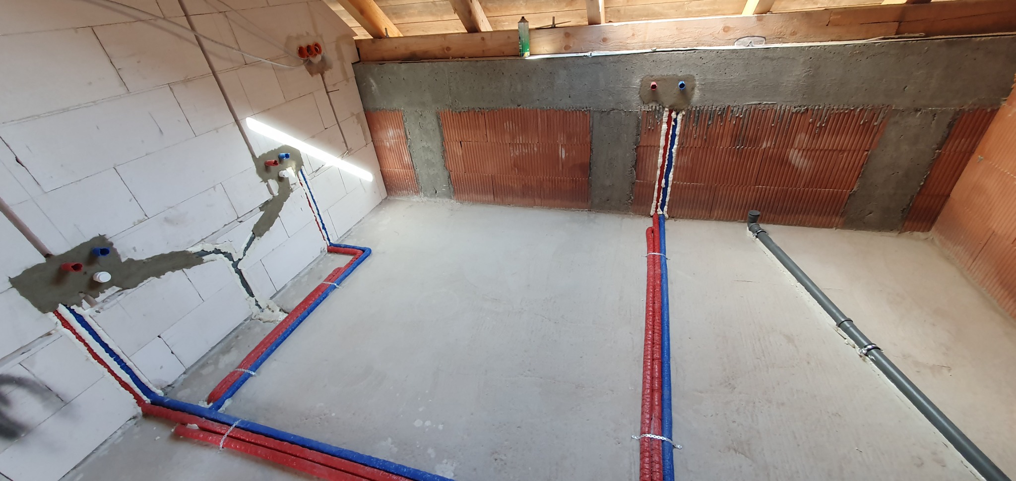 Wnętrze w Wiśniowej w trakcie prac hydraulicznych: widoczne rozprowadzone rury (czerwone i niebieskie) przy ścianach z betonu komórkowego i cegły, częściowo zamurowane i zabezpieczone pianką...