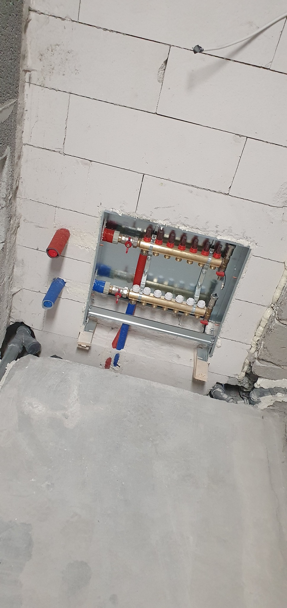 Rozdzielacz ogrzewania podłogowego zamontowany w ścianie z bloczków, widoczne rury PEX w kolorze czerwonym i niebieskim, oraz zawory regulacyjne.