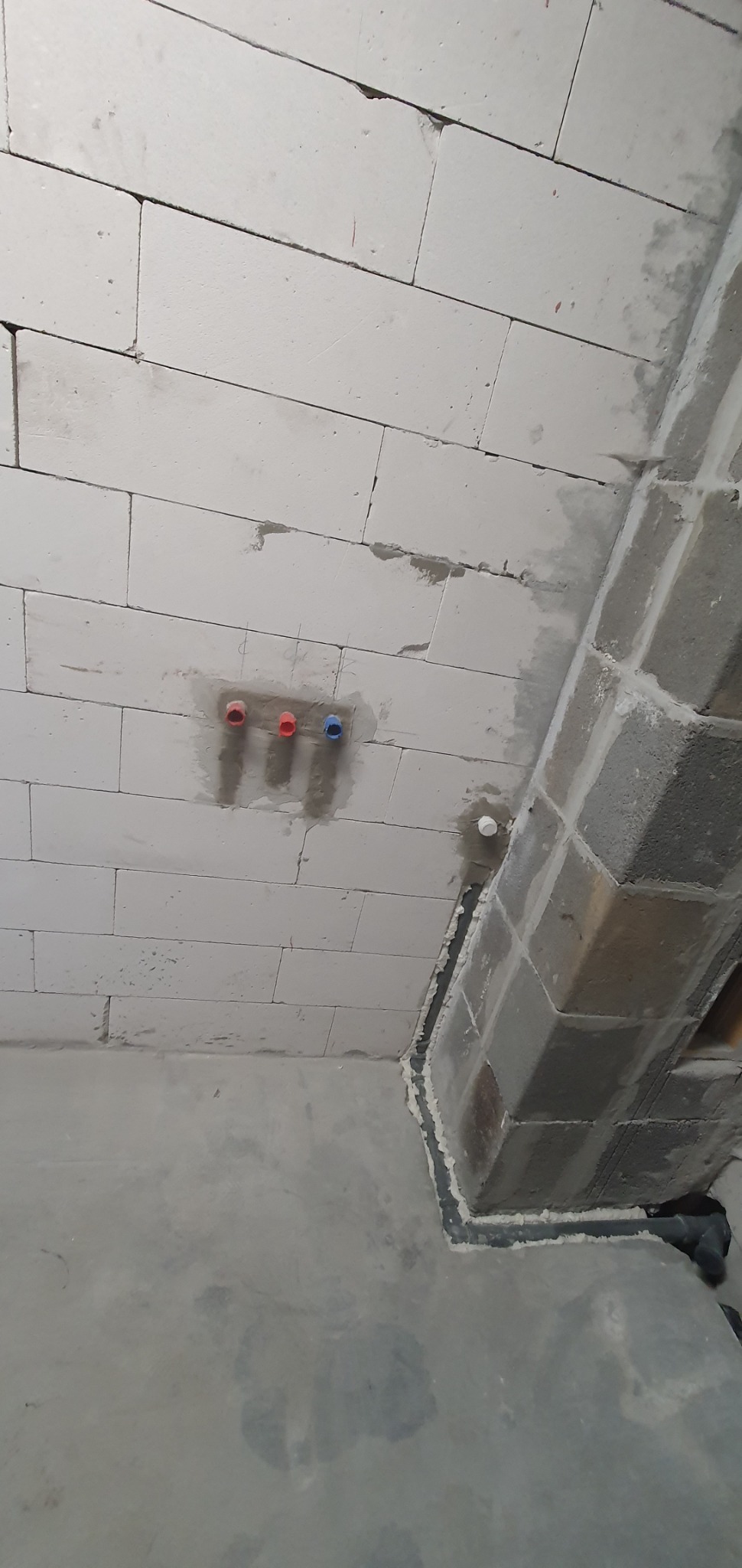 Surowy stan instalacji hydraulicznej w ścianie z betonu komórkowego, widoczne trzy wyprowadzenia rur w kolorach czerwonym i niebieskim, oraz pion instalacyjny zabezpieczony uszczelniaczem.