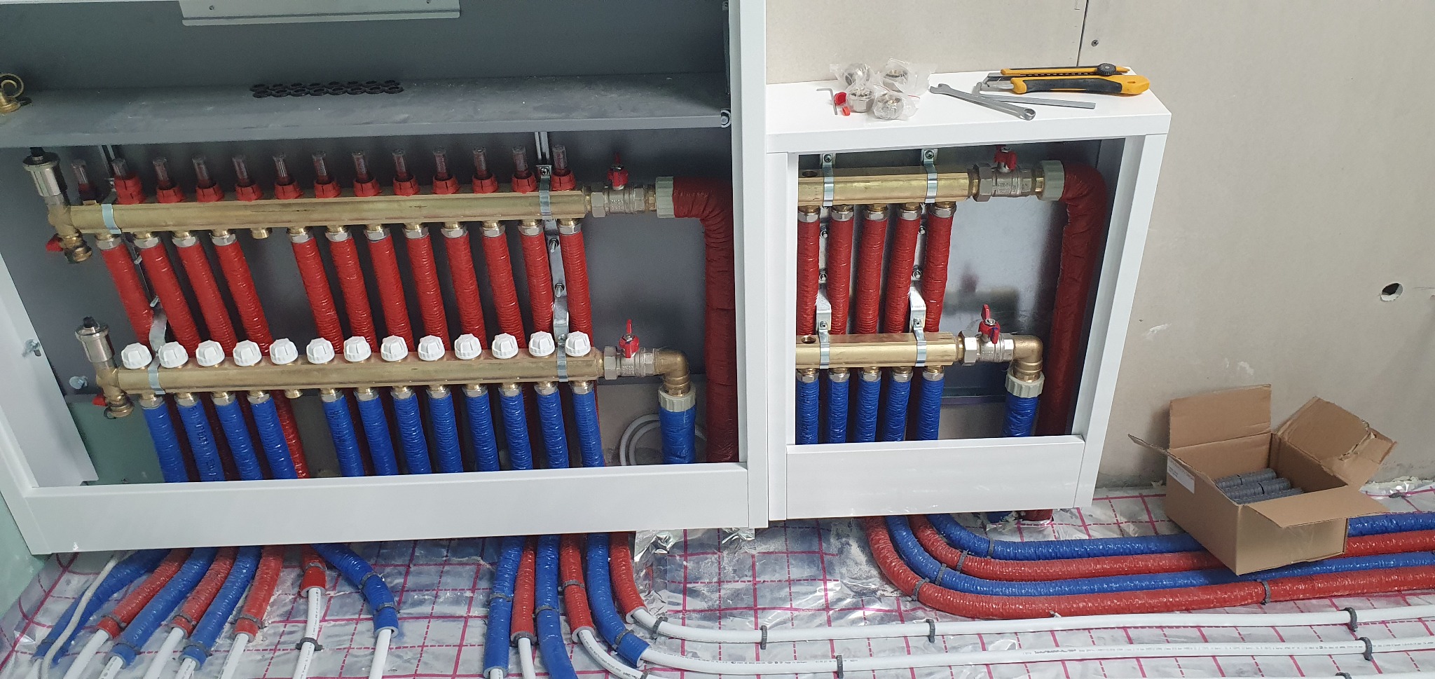 Rozdzielacz ogrzewania podłogowego w szafce, widoczne rury czerwone i niebieskie, narzędzia na półce i karton z elementami montażowymi.