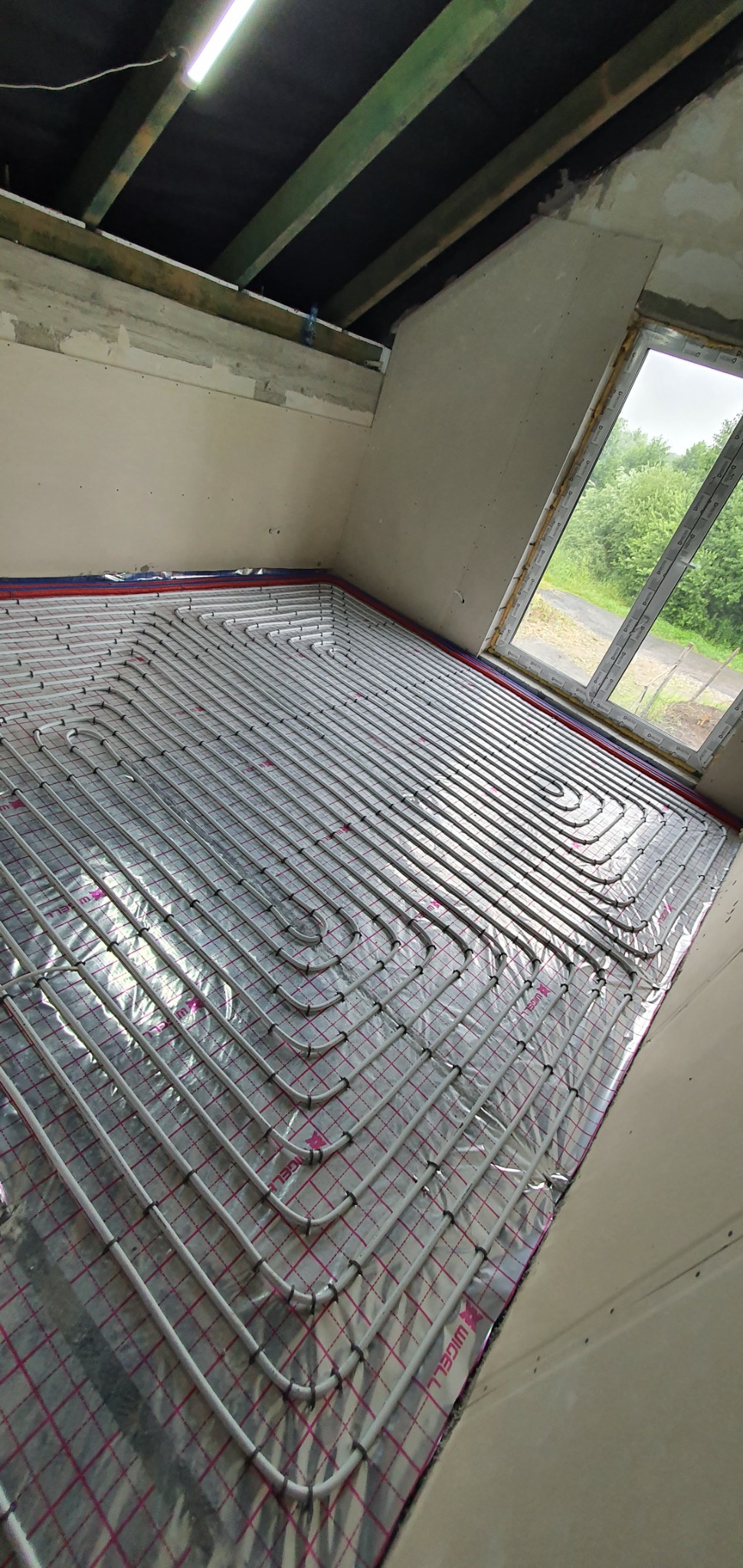 Montaż ogrzewania podłogowego: białe rury ułożone w pętle na srebrnej folii izolacyjnej z czerwonym nadrukiem kratki, widok na okno i surowy stan ścian w trakcie remontu.