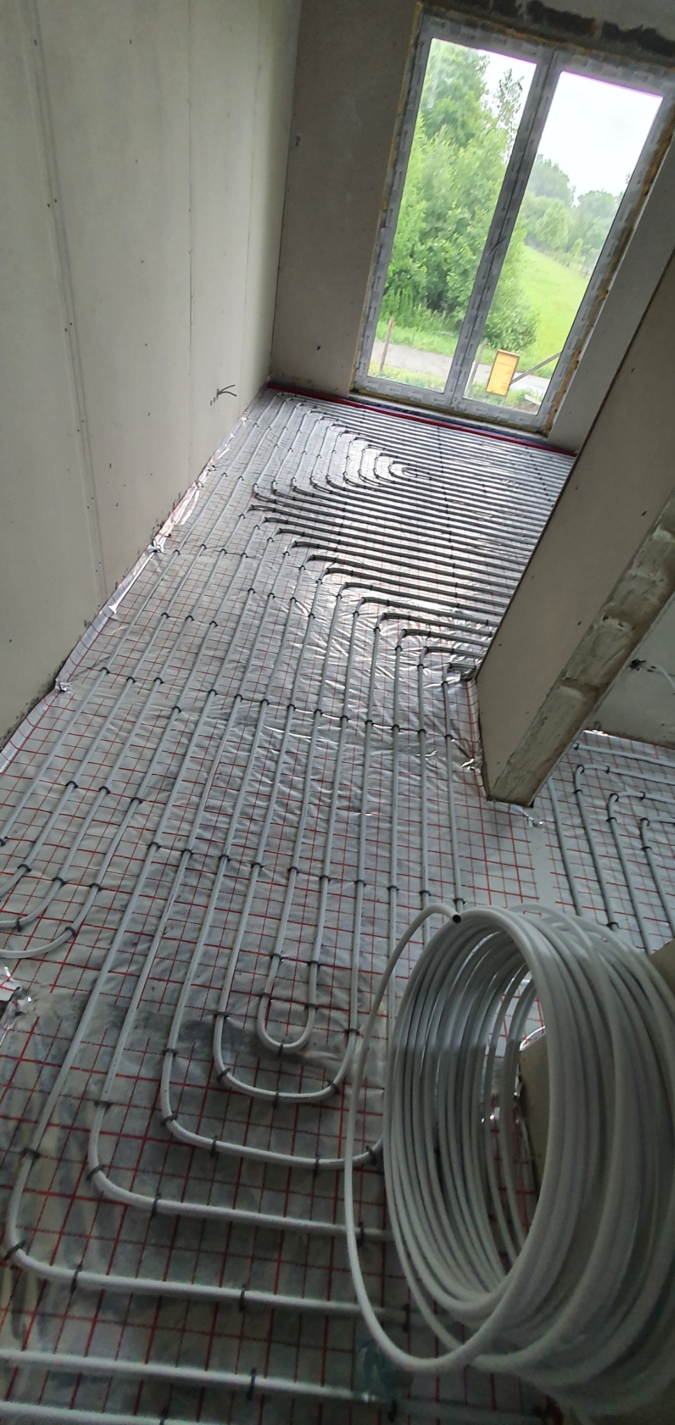 Rozłożone rury ogrzewania podłogowego na folii z siatką, widoczna rolka rur, okno z widokiem na zieleń w tle.