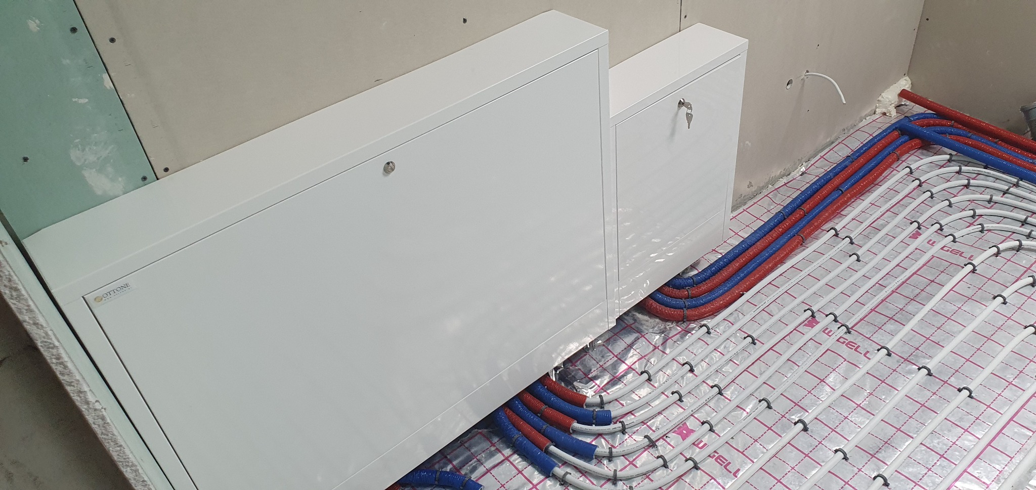 Rozłożone rury ogrzewania podłogowego w folii izolacyjnej, widoczne białe szafki zamykane na klucz i fragment ściany z płyt gipsowo-kartonowych.