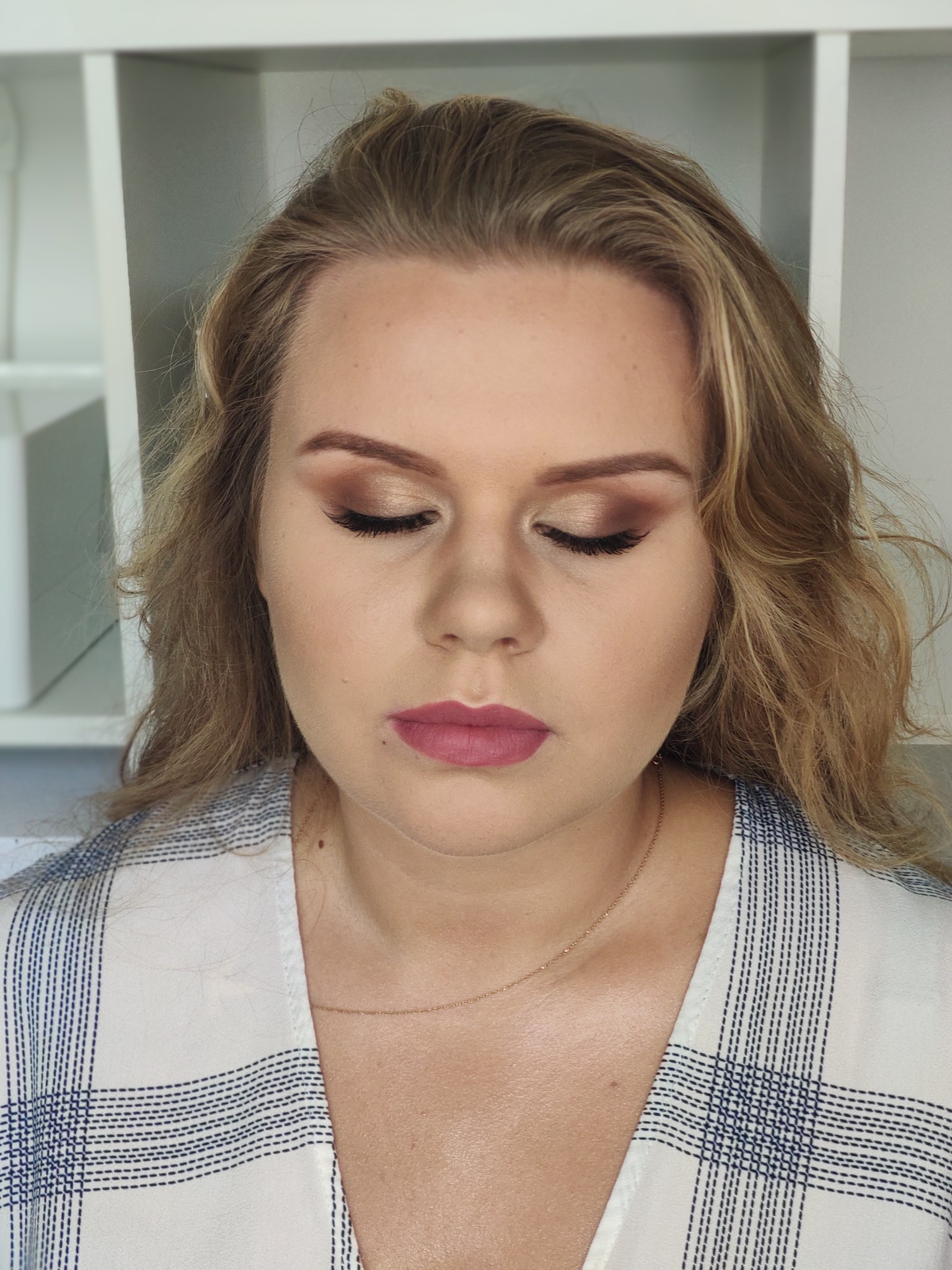 Portret kobiety z zamkniętymi oczami, prezentujący makijaż oczu w odcieniach brązu i złota oraz matowe, różowe usta, z delikatną złotą biżuterią na szyi i bluzką w kratę.