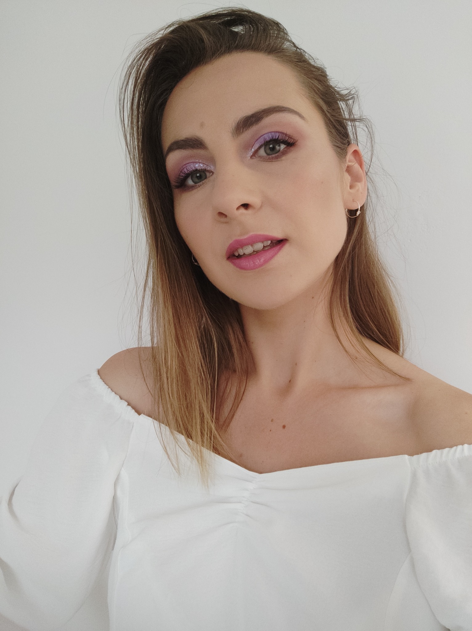 Portret kobiety z profesjonalnym makijażem oczu w odcieniach fioletu i różu, podkreślonym błyszczącym brokatem, oraz różową pomadką na ustach, ubrana w białą bluzkę z odkrytymi ramionami.