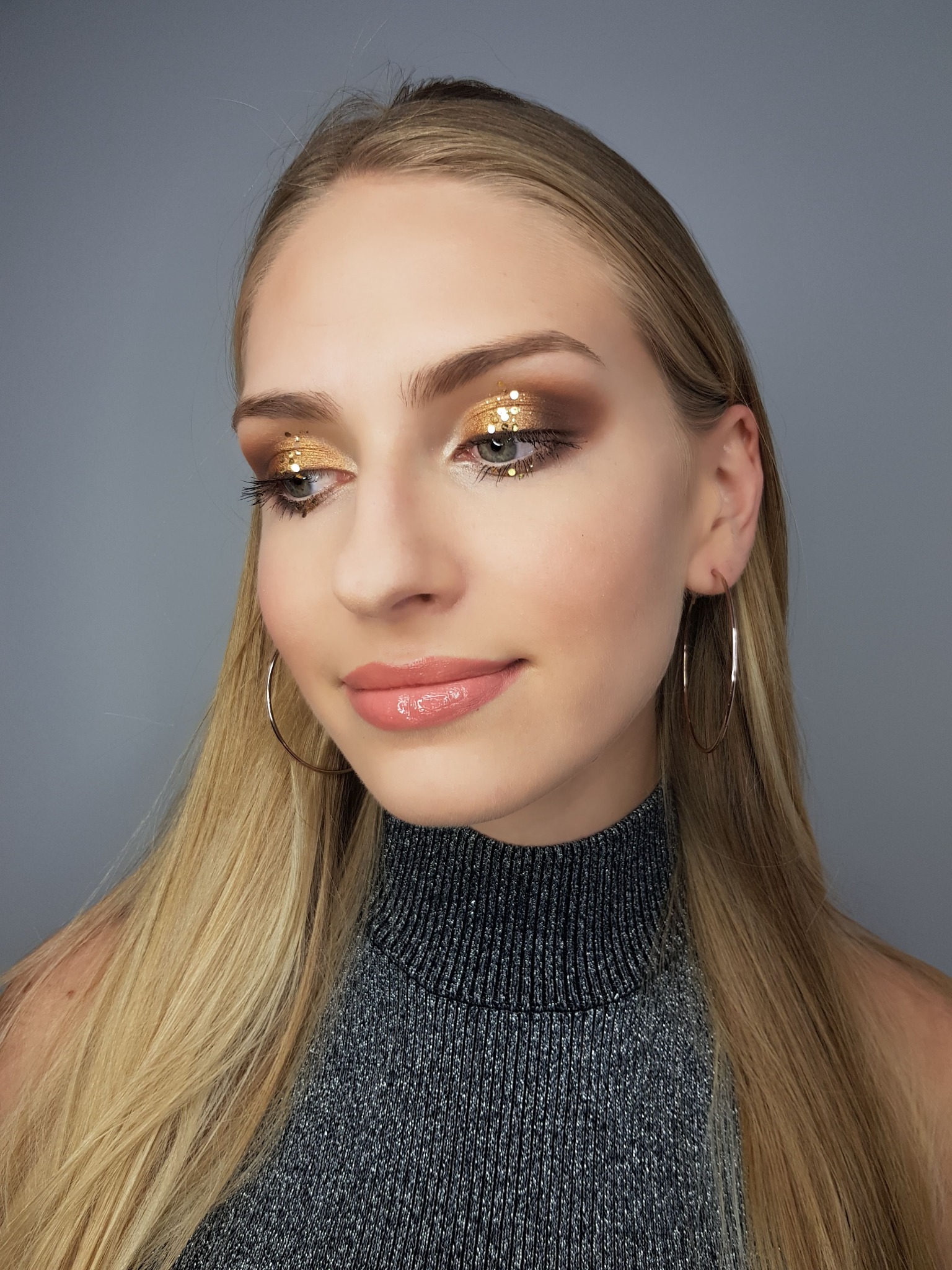 Portret kobiety z blond włosami i błyszczącym, złotym makijażem oczu oraz błyszczykiem na ustach, pozującej na szarym tle.