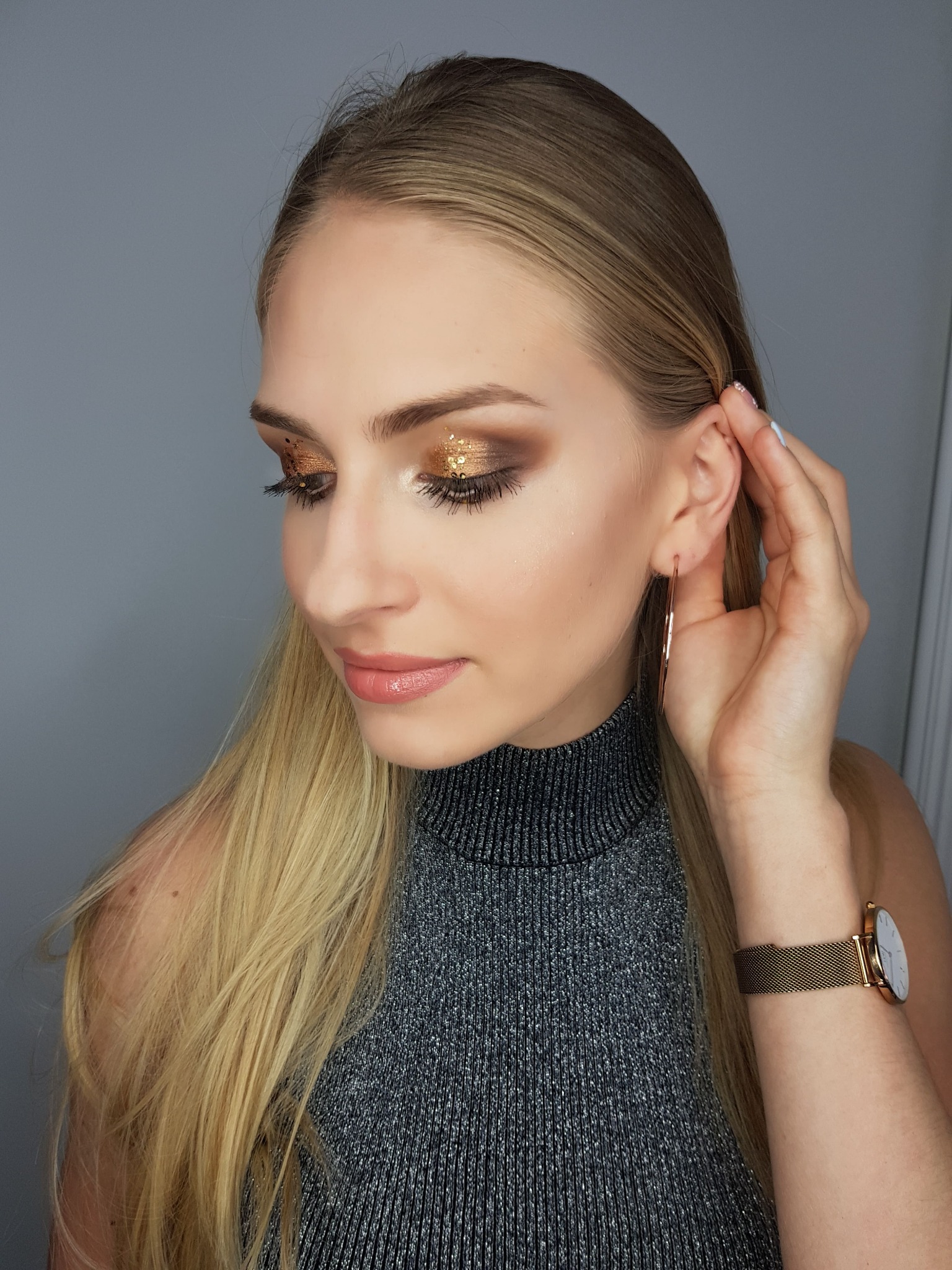 Portret kobiety z blond włosami, w srebrnej bluzce, z wieczorowym makijażem oczu z brokatem, dotykającej ucha z długim, złotym kolczykiem i zegarkiem na ręce.