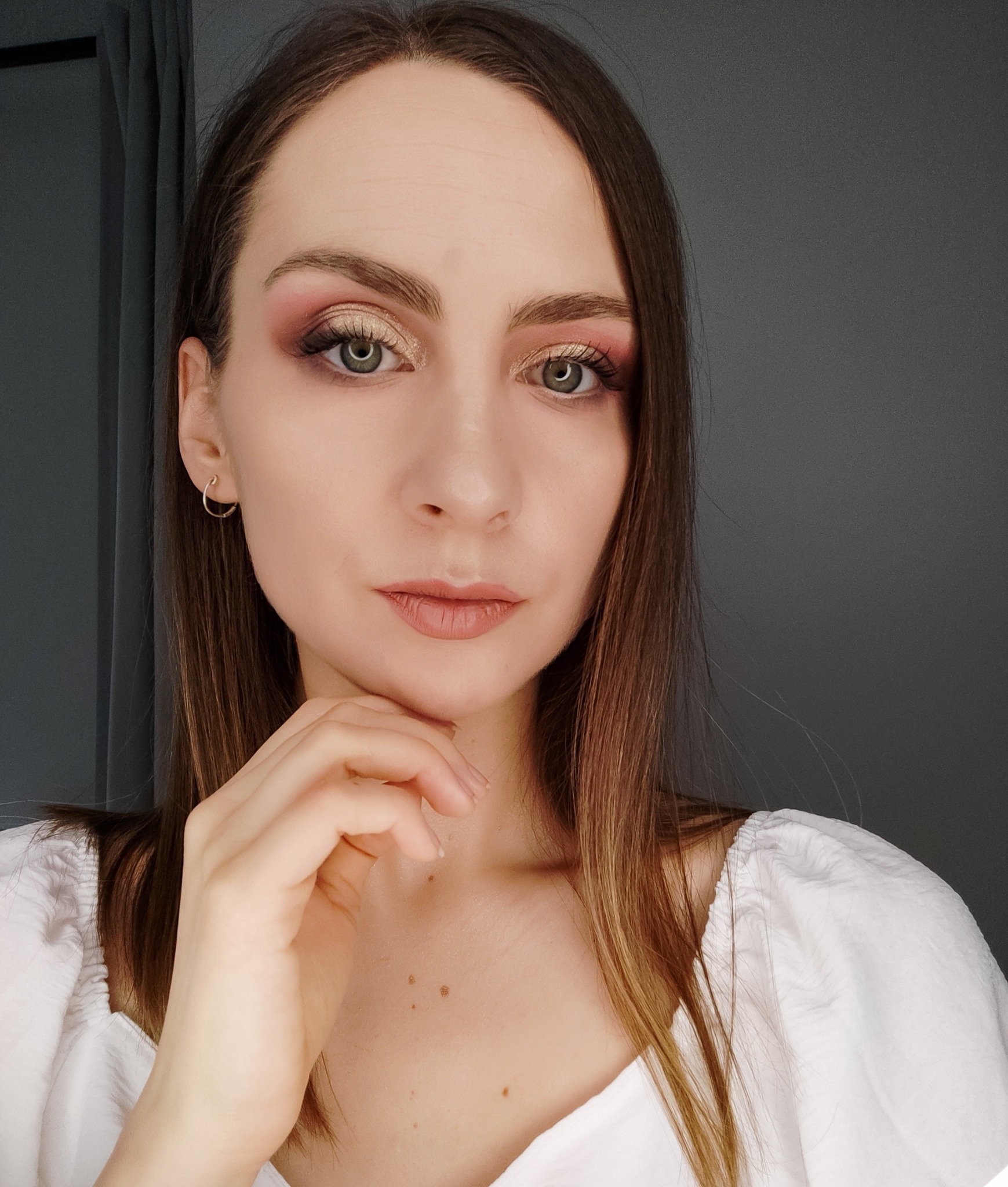 Portret kobiety z subtelnym makijażem oczu w odcieniach różu i złota, lekko pochyloną głową i dłonią pod brodą, na szarym tle.