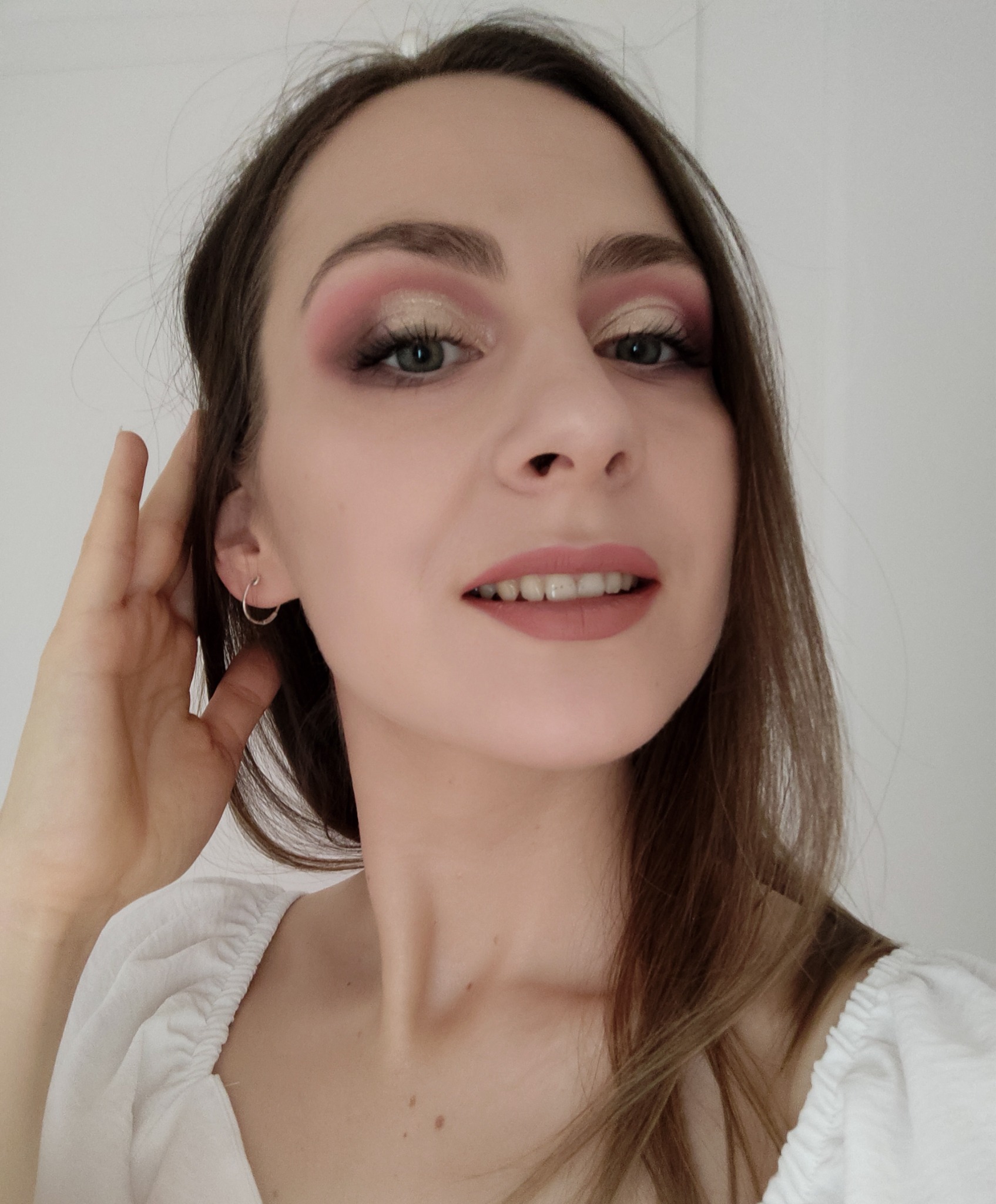 Portret kobiety z delikatnym makijażem oczu w odcieniach różu i złota, oraz pomadką w kolorze nude; widoczna srebrna kolczyk w uchu i dłoń przy twarzy.