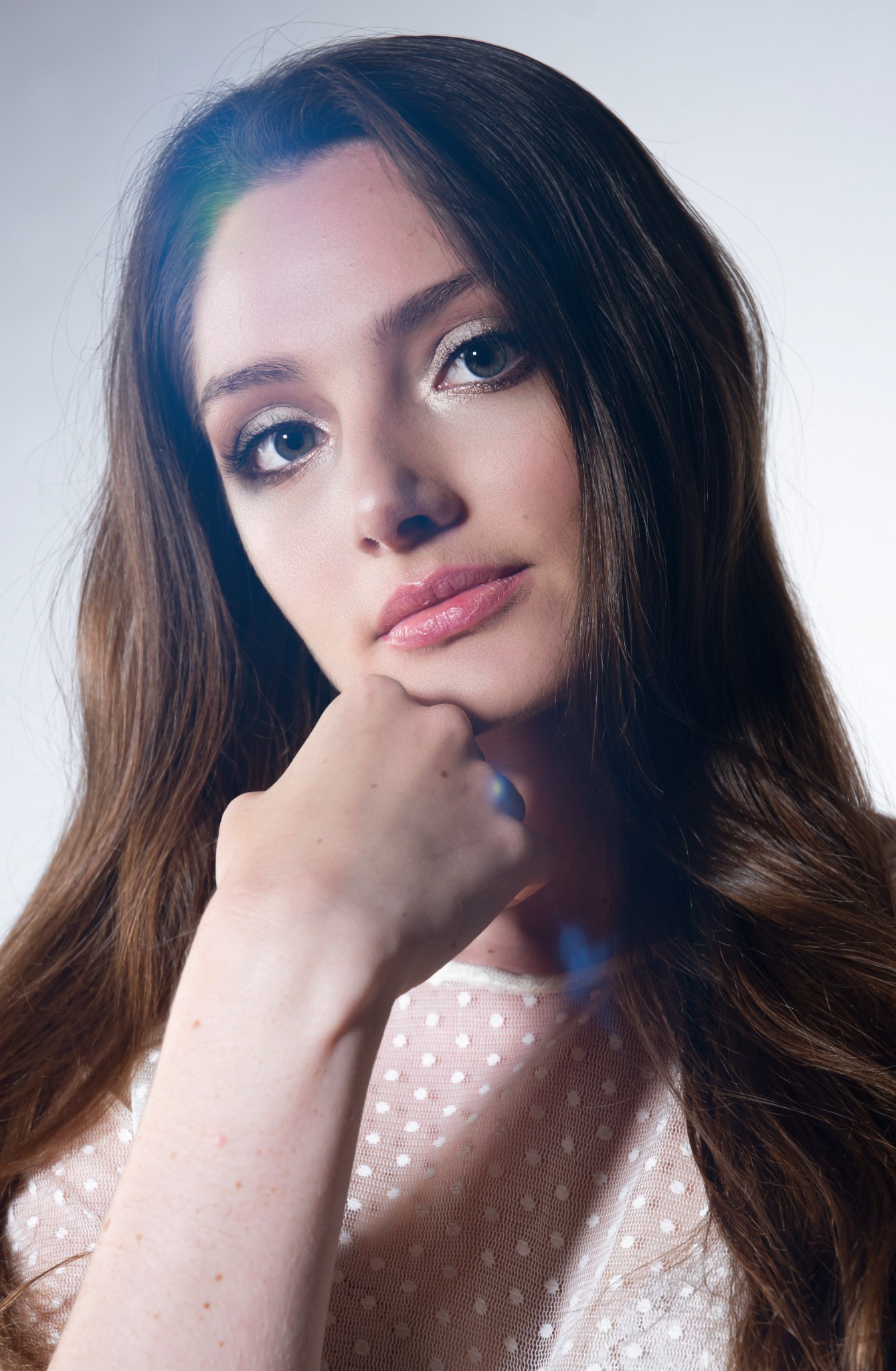 Portret kobiety z długimi brązowymi włosami, opierającej podbródek na dłoni, z delikatnym makijażem oczu i błyszczącymi ustami, ubrana w bluzkę w białe kropki na jasnym tle.