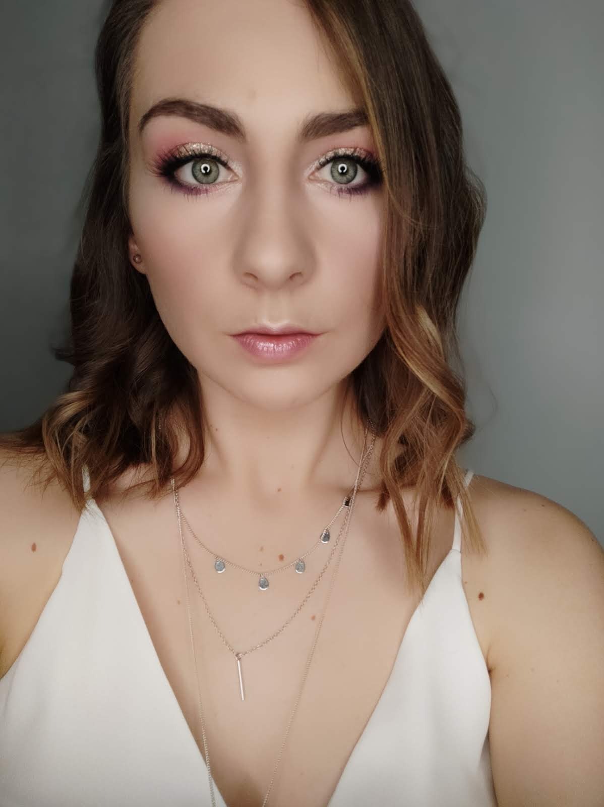 Portret kobiety z delikatnym makijażem oczu w odcieniach różu i złota, noszącej srebrny naszyjnik z zawieszkami na tle szarej ściany.