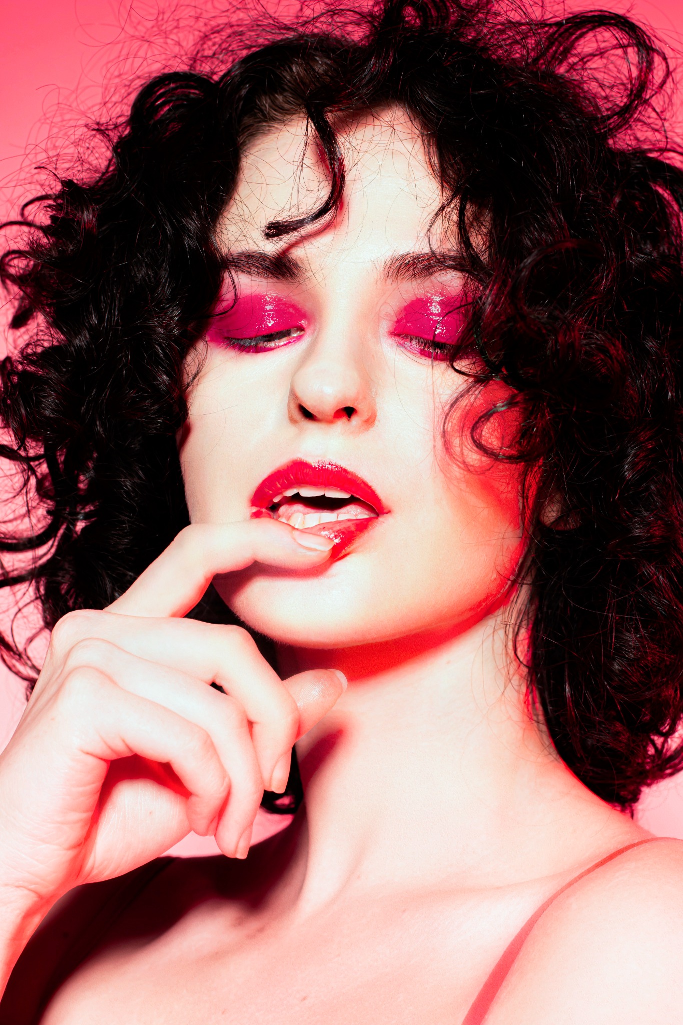 Portret kobiety z ciemnymi kręconymi włosami, intensywnym różowym makijażem oczu i ust, palec delikatnie dotyka ust, całość w różowej tonacji.