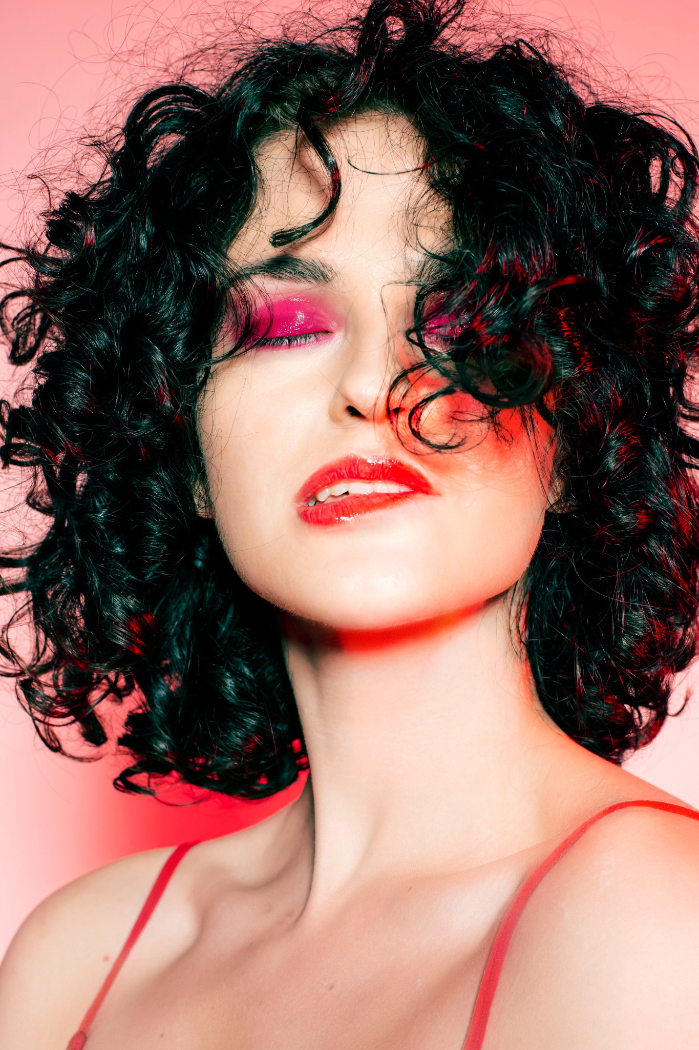Portret kobiety z kręconymi włosami i intensywnym makijażem oczu i ust w Krakowie, na różowym tle.
