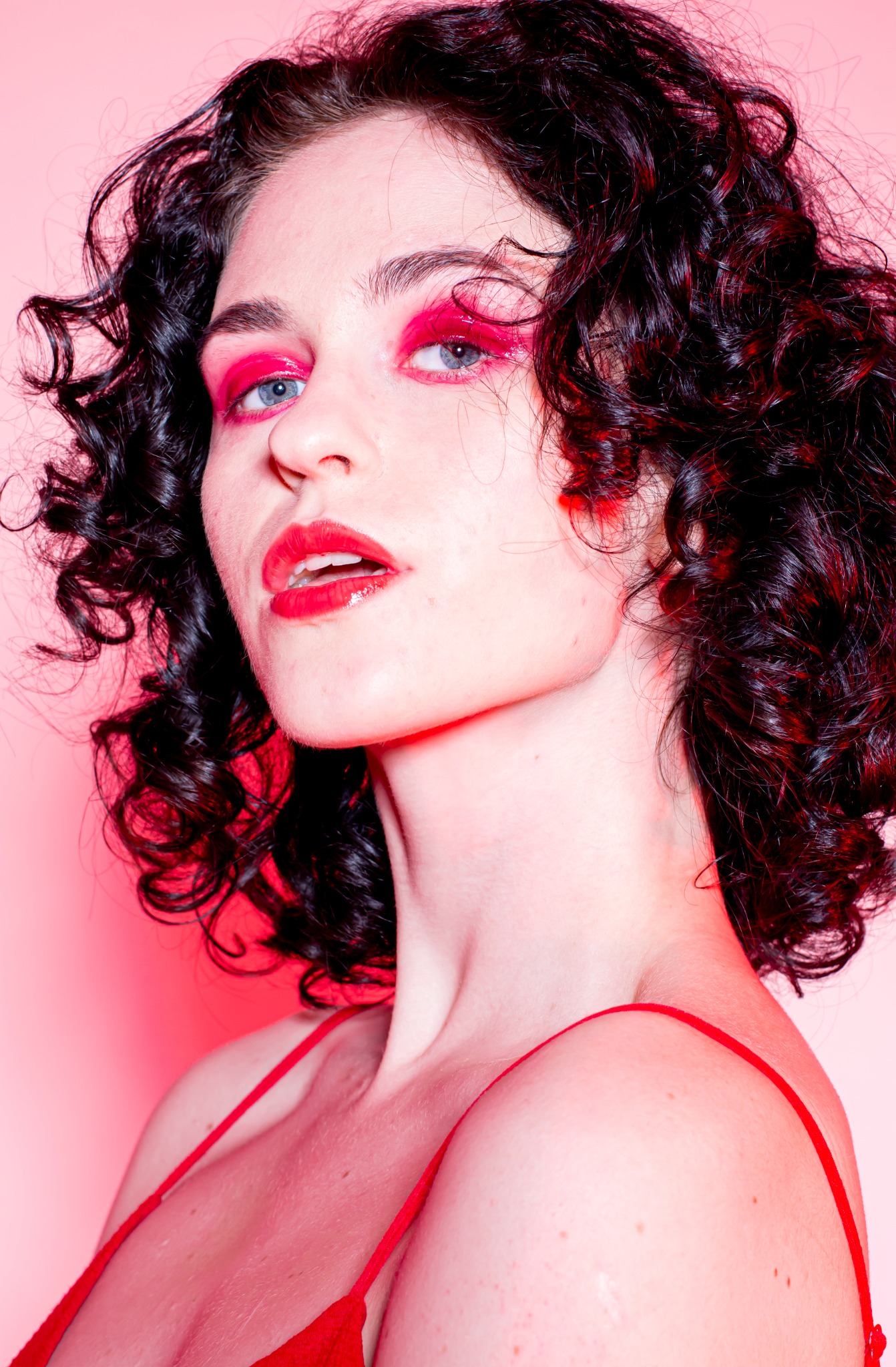 Portret kobiety z intensywnym makijażem w odcieniach czerwieni, kręcone ciemne włosy, skierowana w lewo, na jednolitym różowym tle.