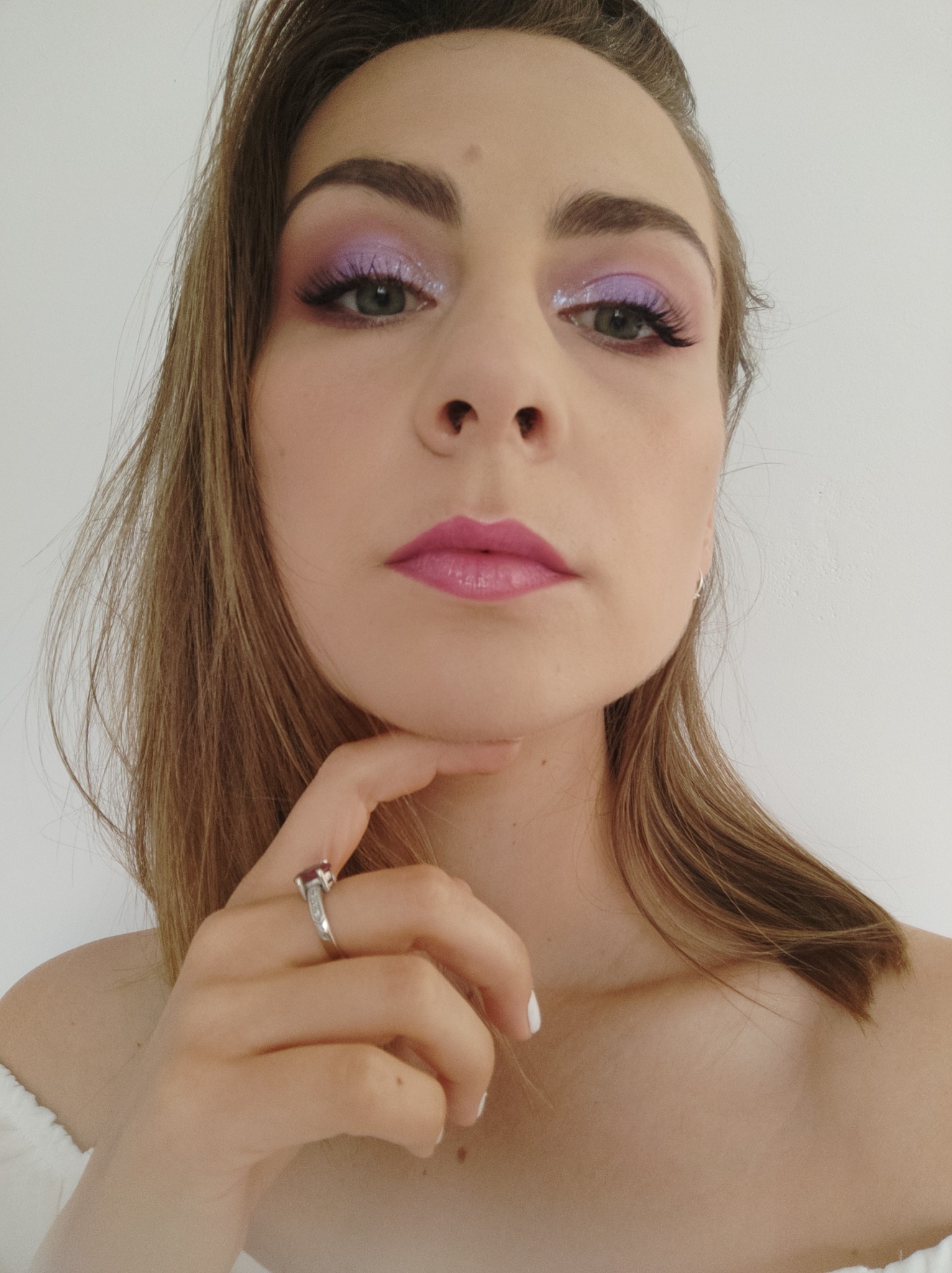 Portret kobiety z profesjonalnym makijażem oczu w odcieniach fioletu i różowe usta, dłoń z pierścionkiem podparta pod brodą, skierowana w stronę obiektywu.